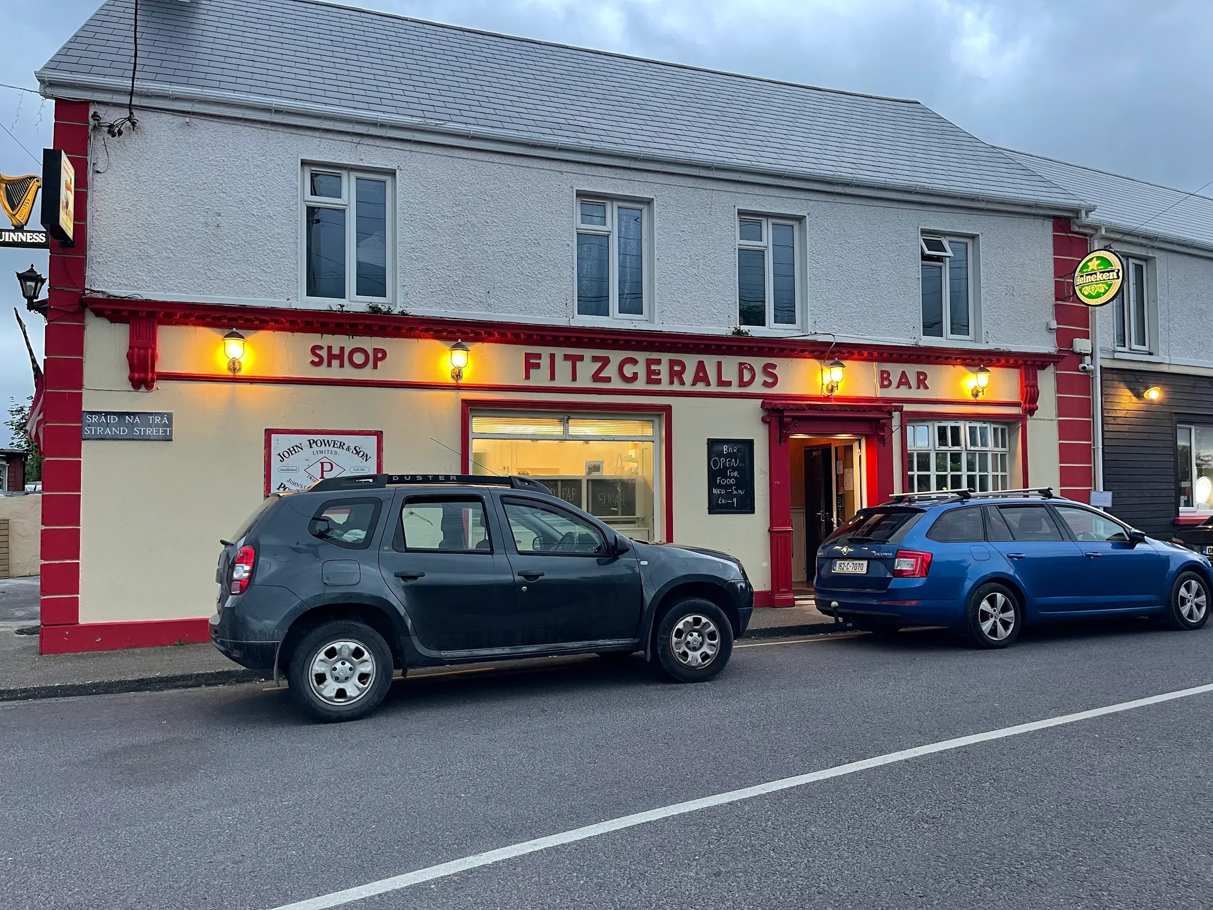 The exterior of Fitzgeralds Bar and shop with two cars parked in front, illuminated yellow lights, and a Heineken sign on the right side.