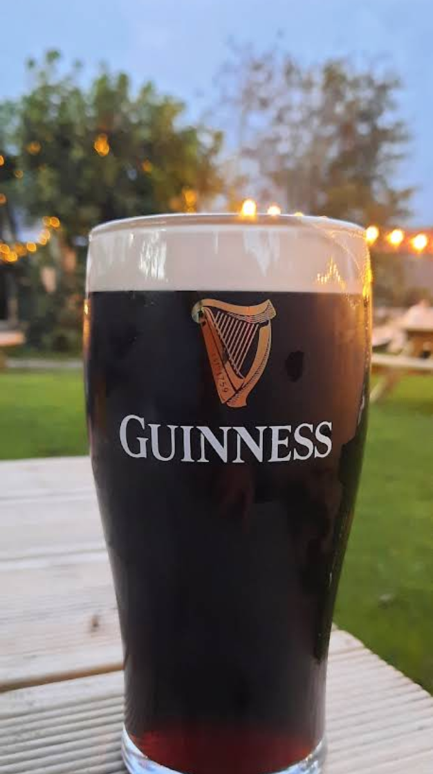 A glass of Guinness stout beer with a foamy head on a wooden table outdoors during evening, with trees and string lights in the background.