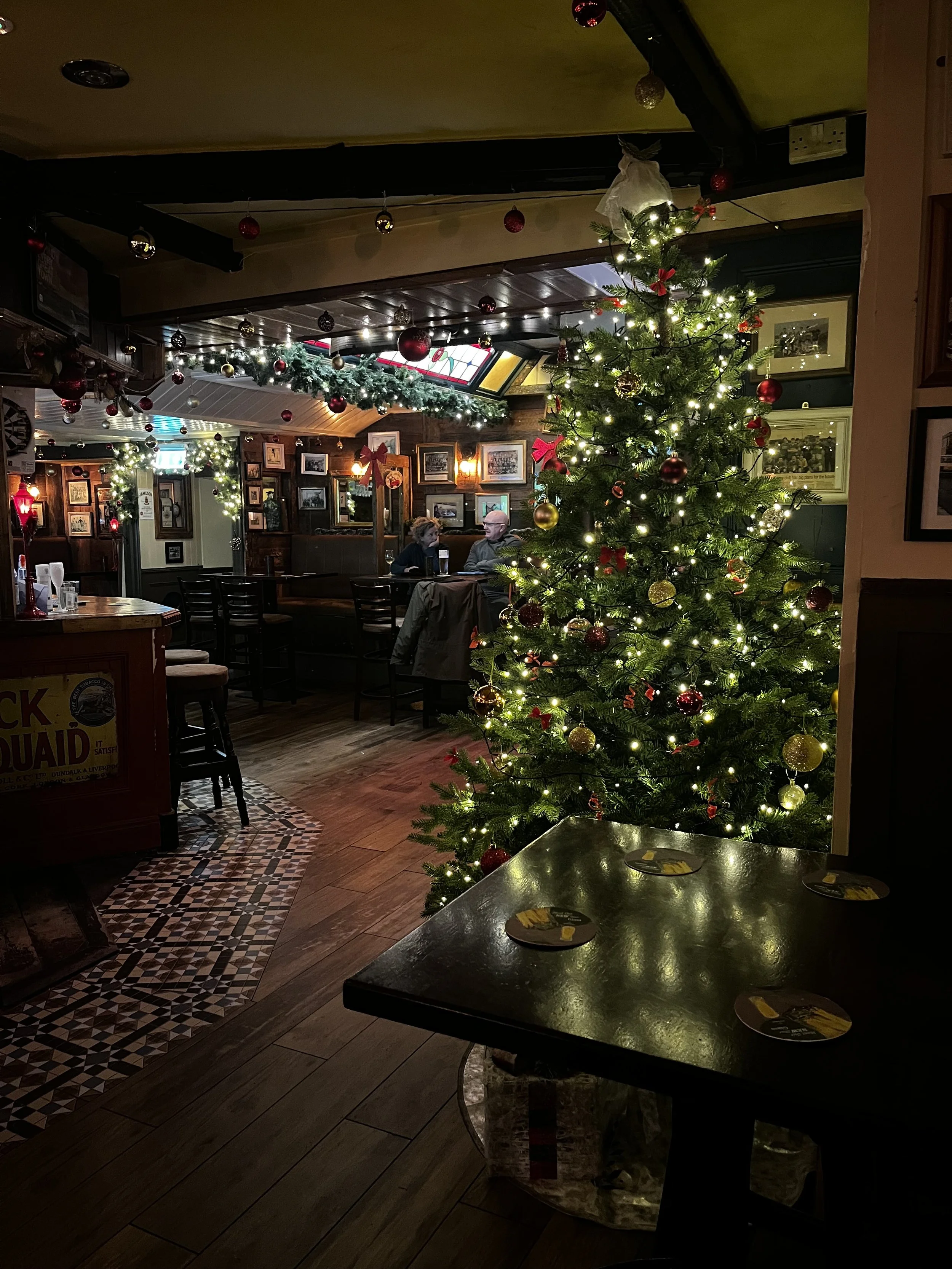 Decorated Christmas tree with lights and red and gold ornaments inside a cozy pub with wooden furniture, framed pictures on the walls, and people sitting at tables.