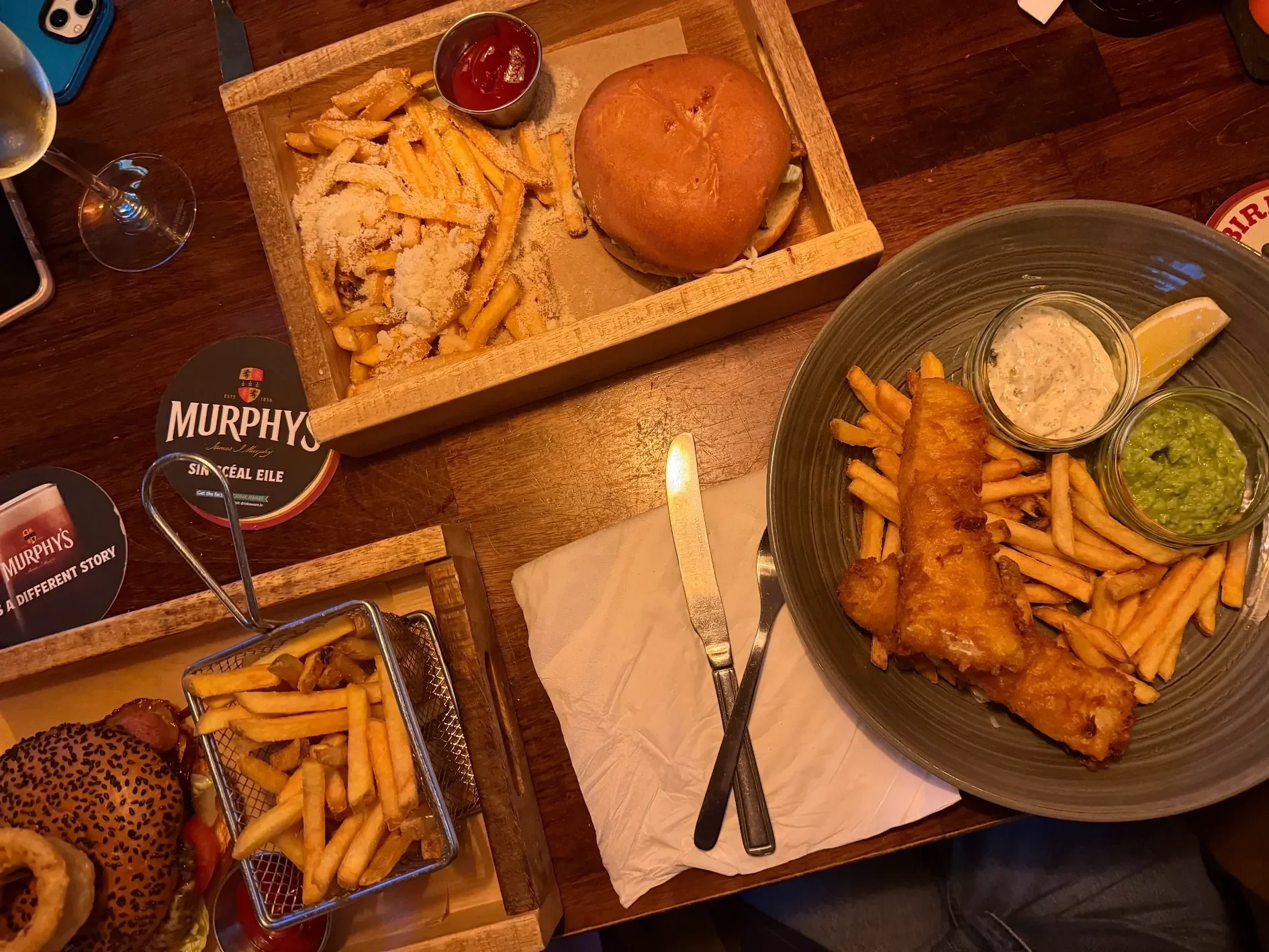 A meal with fried fish, French fries, and condiments on a large plate, a burger with fries in a wooden tray, and a small basket of fries with a burger on a wooden table.