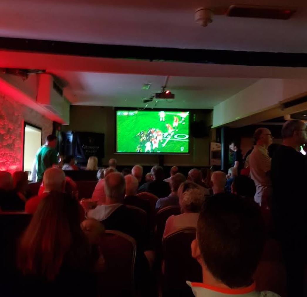 A group of people seated in a dark room watching a football game on a large screen, with some people standing and talking.