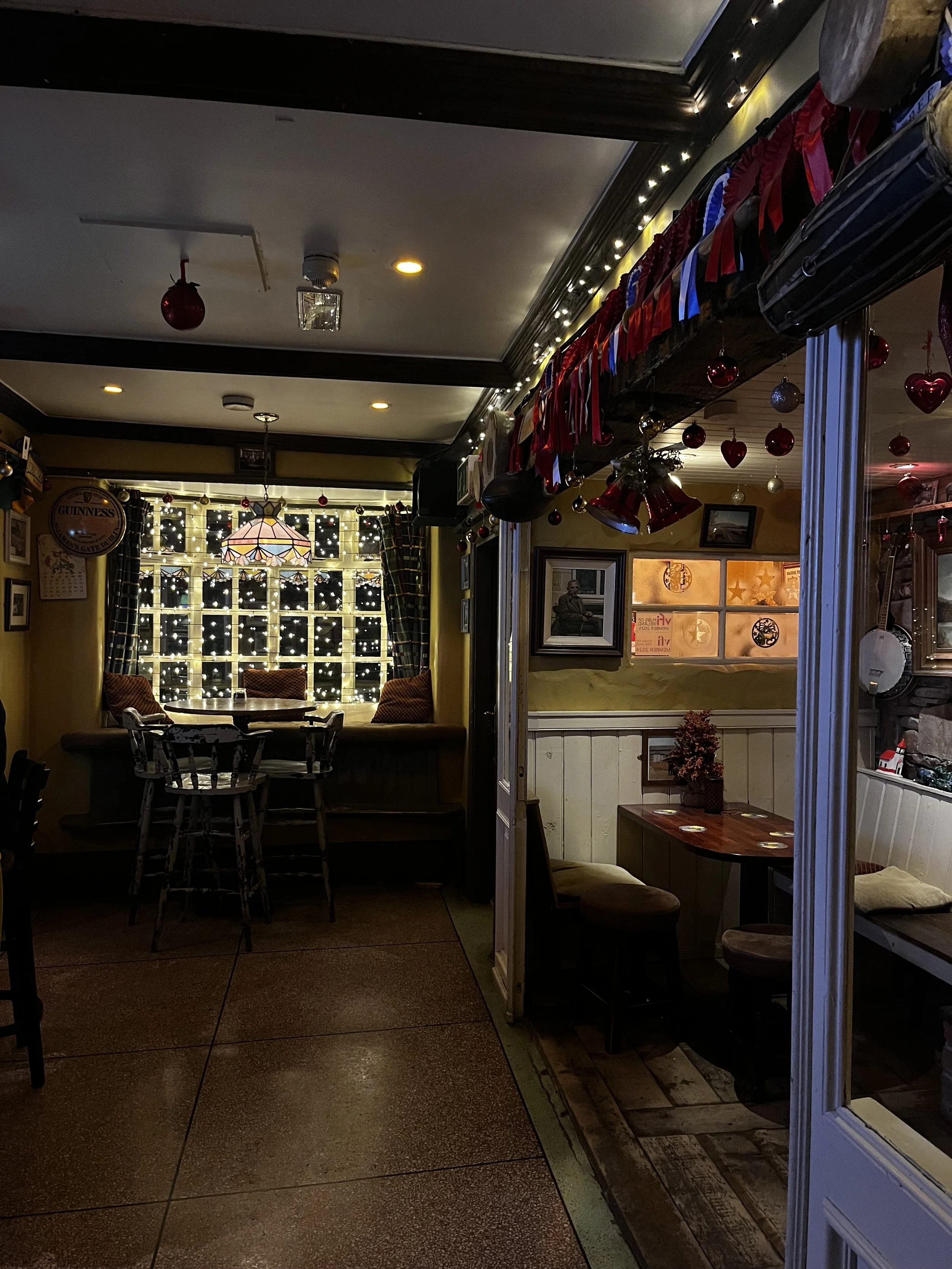 Interior view of a cozy restaurant with holiday decorations, including red and silver heart-shaped ornaments and ribbons hanging from the ceiling. There are tables and chairs, with a window adorned with string lights and curtains in the background.