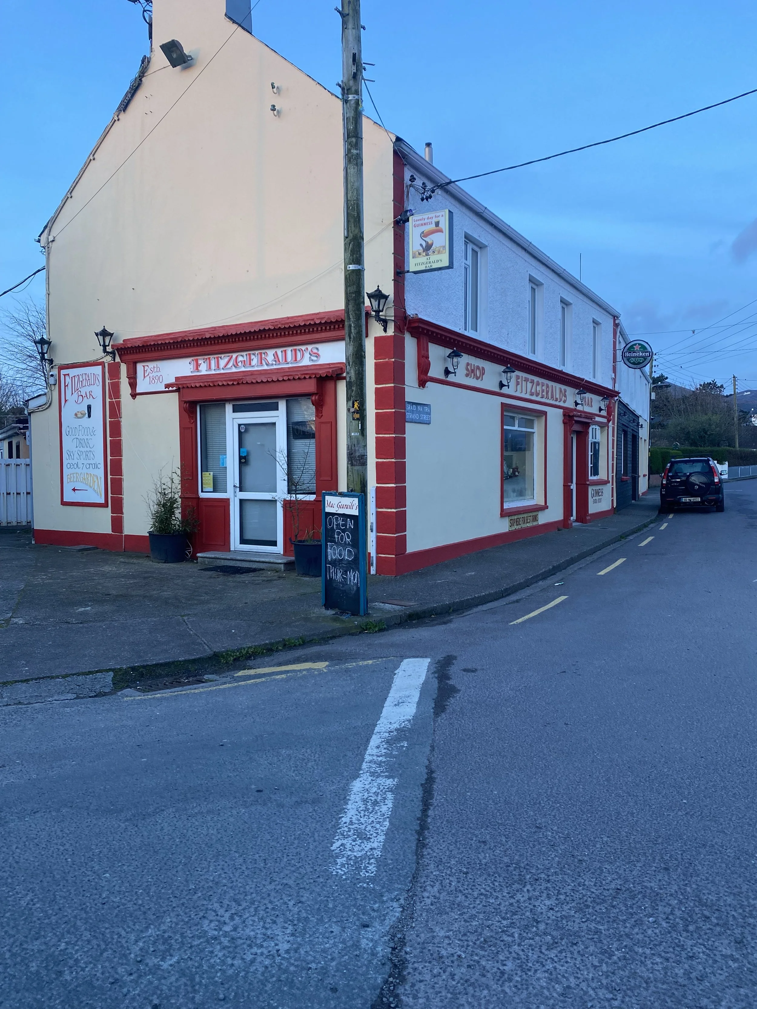 Corner view of Fitzgerald's bar and shop in a small town, with a yellow building featuring red trim, a door with a small window, a sidewalk, and a parking area with a black car. There are signs indicating food and drinks, and a chalkboard sign outsid