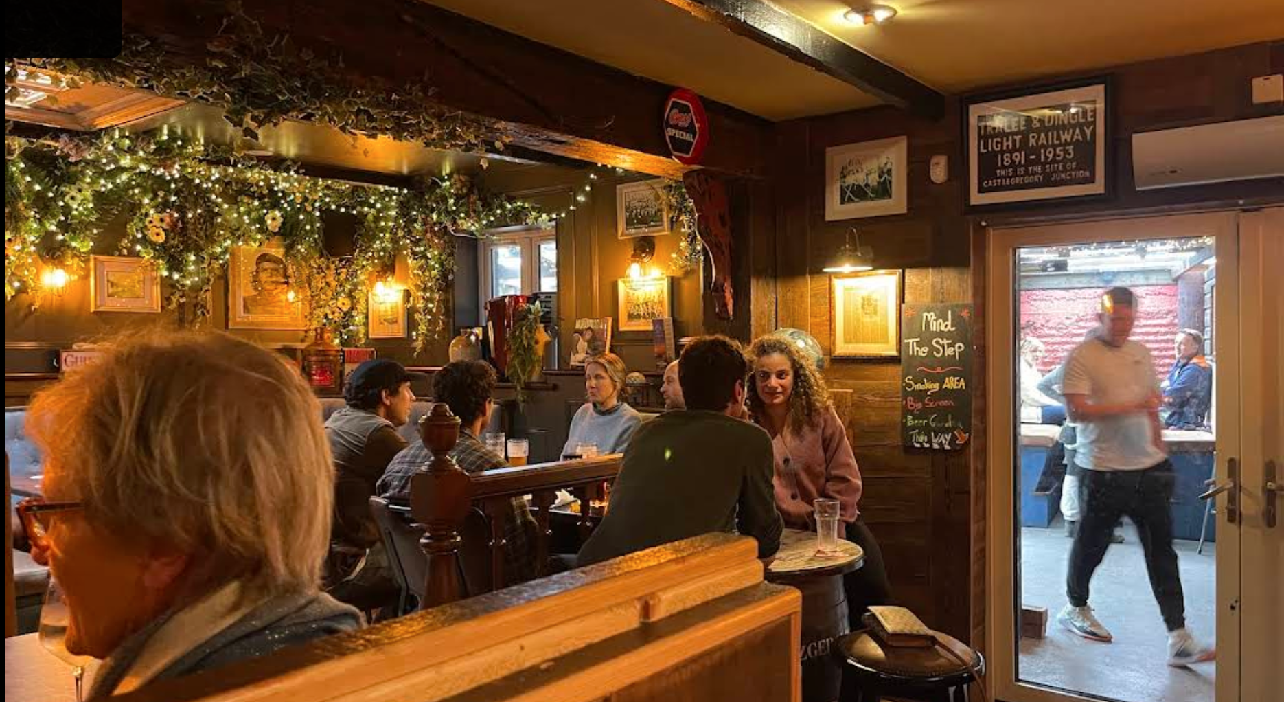 People dining inside a cozy restaurant with wooden walls, framed pictures, and warm lighting. Outside through the glass door, a man walks by while others sit at an outdoor patio.