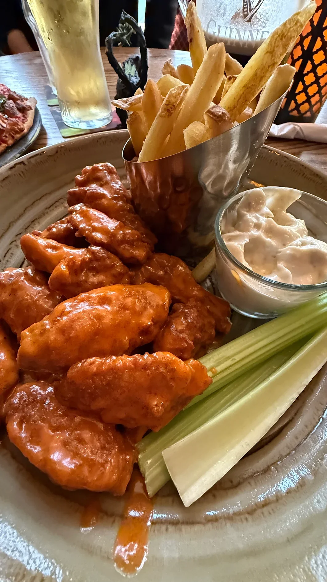 Plate of buffalo chicken wings with celery sticks, French fries in a metal holder, a bowl of ranch dipping sauce, and a glass of beer.