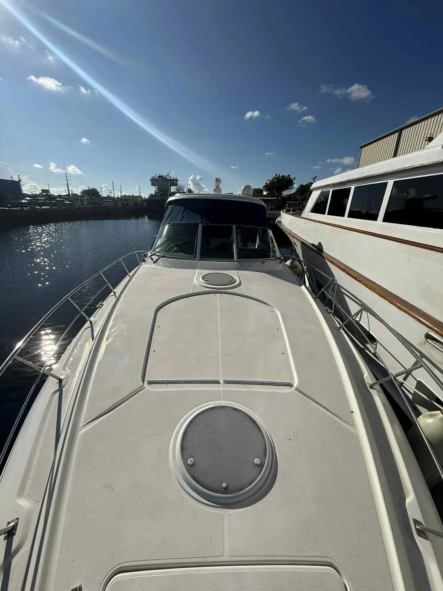 View from the bow of a white yacht docked in a marina, with another yacht to the right and a body of water reflecting the sky. A partly cloudy sky with the sun shining brightly.