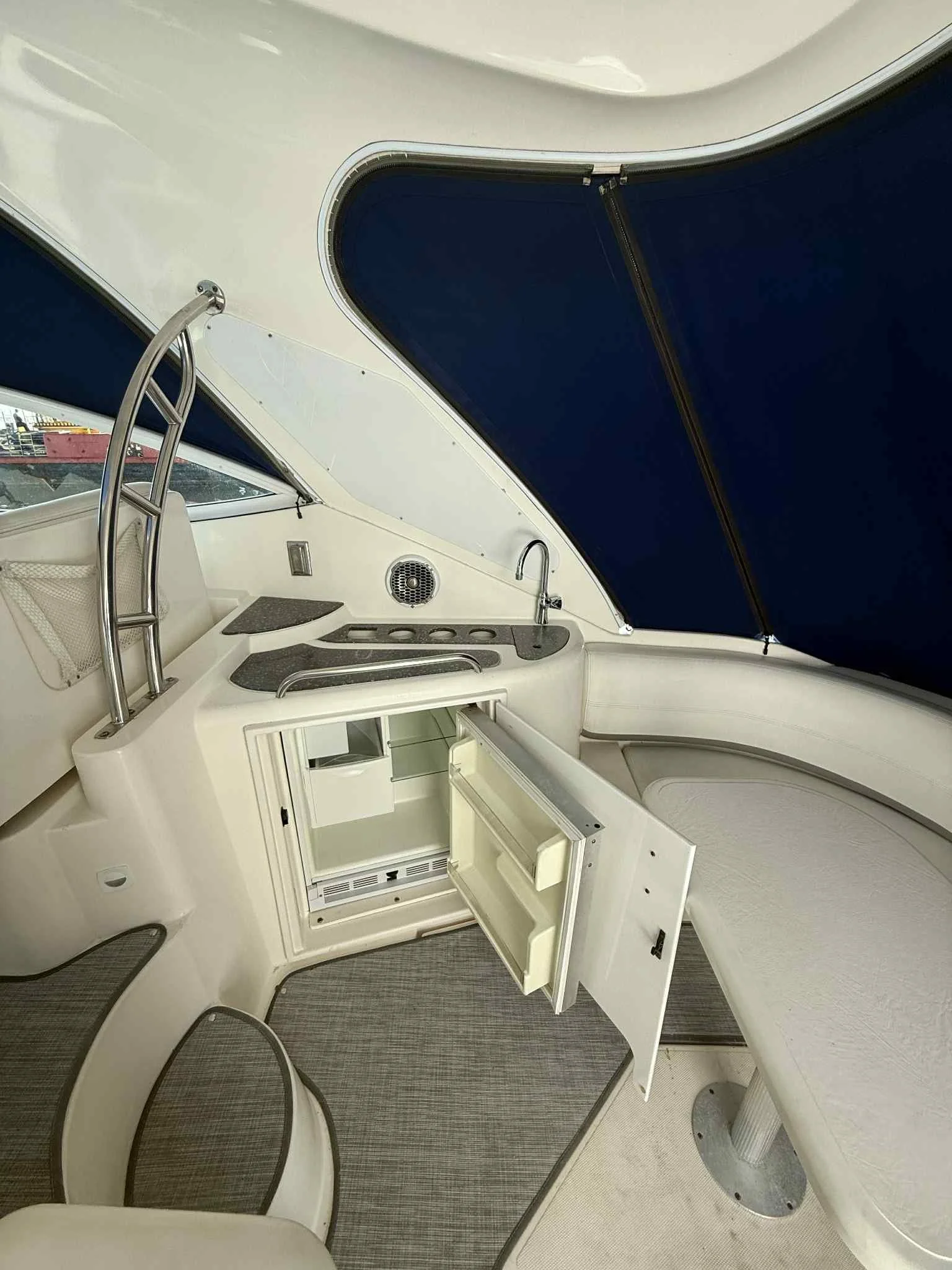 Interior of a boat with a small kitchen area, open mini refrigerator, sink, stove, and seating area under a navy blue canopy.