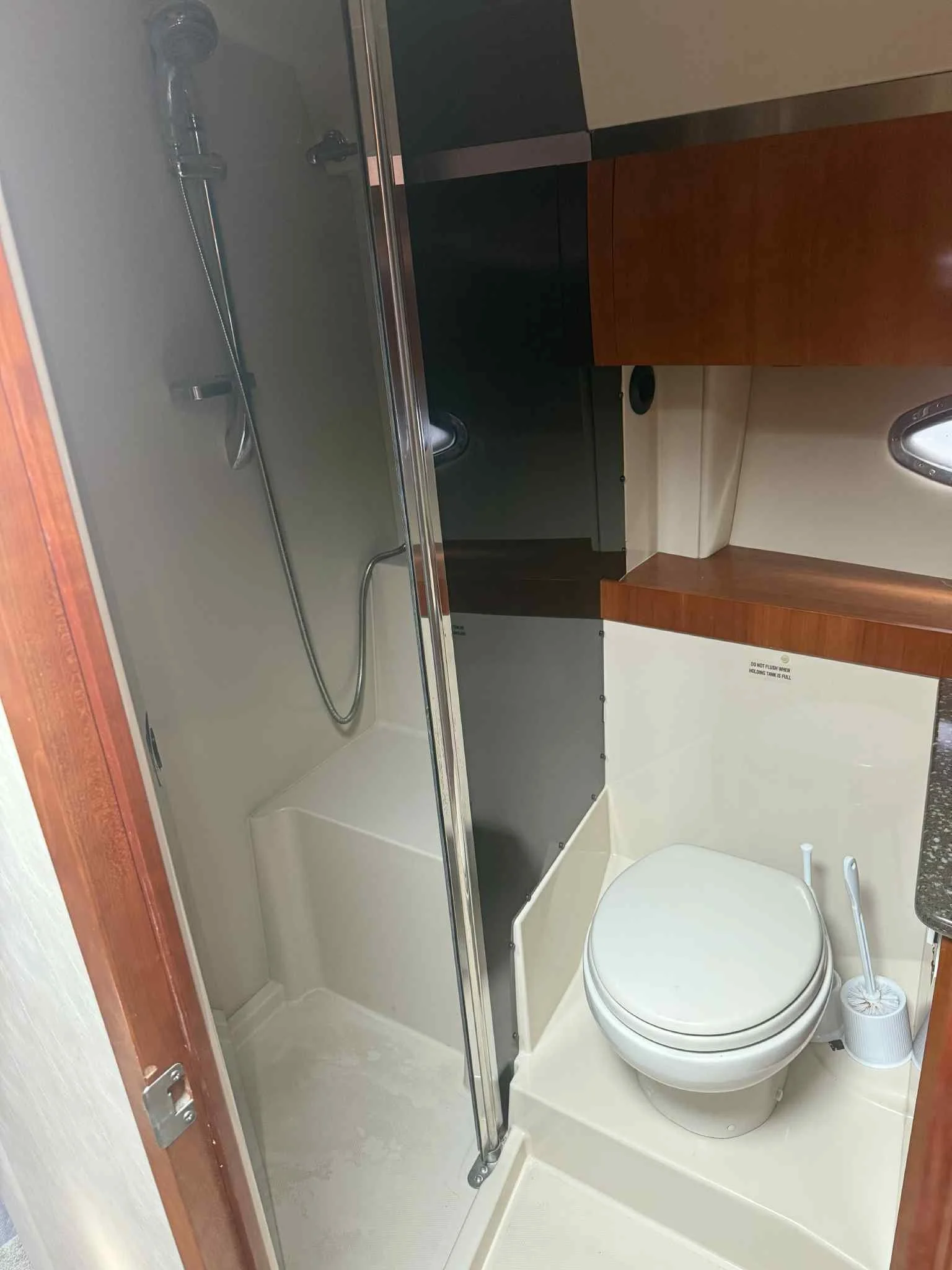 Small bathroom with a shower area, toilet, and wooden cabinets. The shower has a glass door and a silver showerhead with a flexible hose. The toilet is white with a closed lid, and there is a toilet brush in a holder beside it. The upper part of the