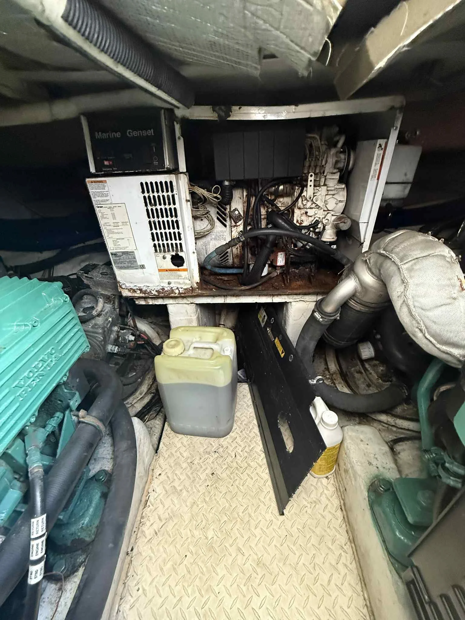 View of a marine engine room with various mechanical and electrical components, including a generator, hoses, and a yellow coolant reservoir.