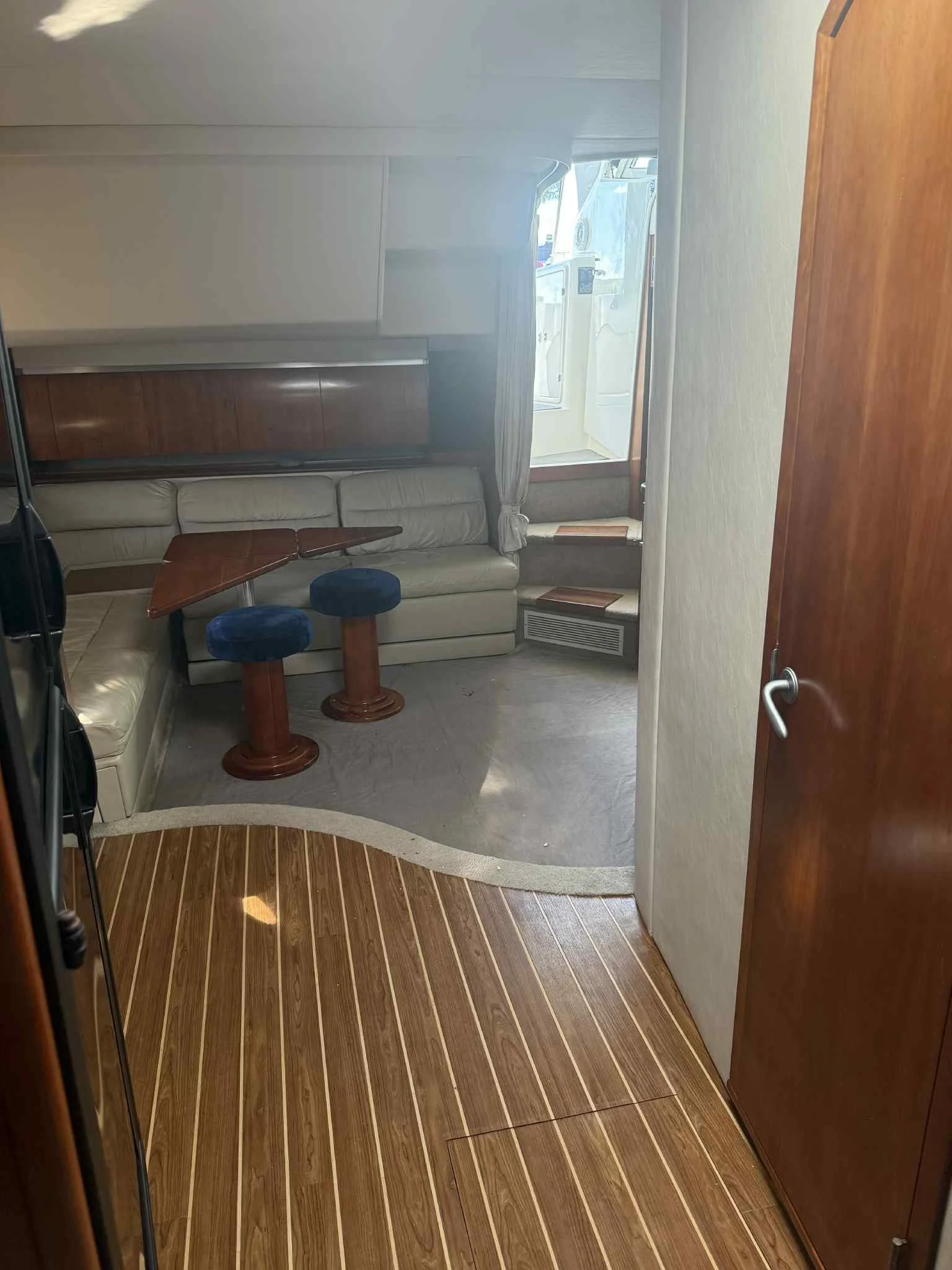 Interior of a boat or yacht with a small seating area featuring a beige bench, two small blue stools, a wooden table, and a staircase leading to an outdoor deck with washing machine and sunlight.
