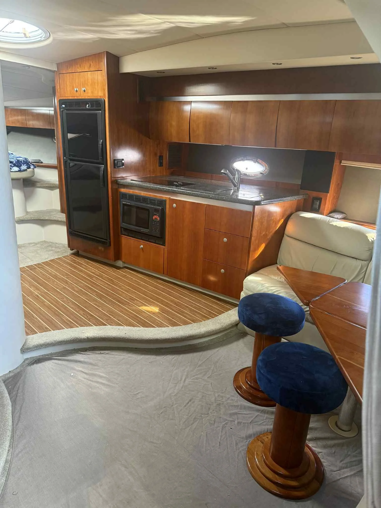 Inside a yacht, featuring a small kitchen with cherry wood cabinets, a microwave, a small sink, and a black refrigerator. Adjacent is a sitting area with a cream leather seat, a wooden table, and two blue upholstered stools. The interior has beige ca