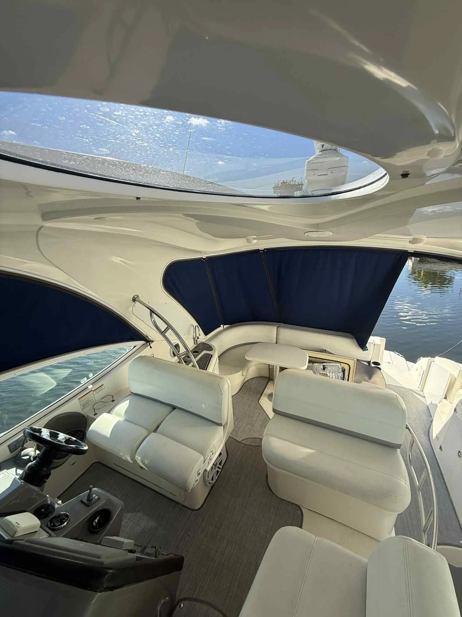 Interior view of a yacht's seating area with white cushioned seats, steering wheel, and a large window, with water and sky visible through a sunroof.