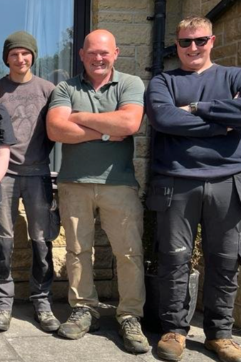 Three men standing outside against a stone wall, smiling with arms crossed or relaxed.