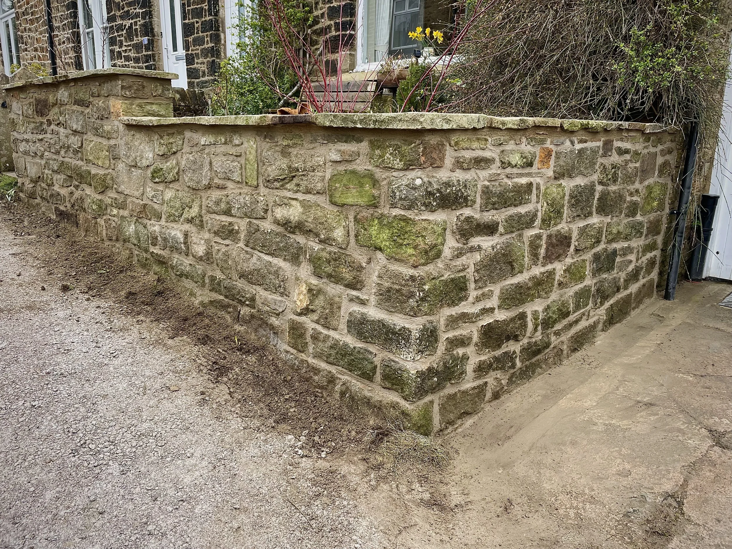 A stone brick garden wall with moss and dirt at its base, with some plants and yellow flowers behind it, situated in front of a house with brick and white siding.