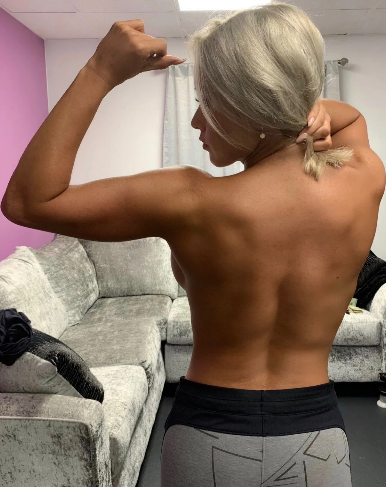 Woman with a recent spray tan from 24 Seven Glow flexing her bicep muscle in a living room with a pink wall and a gray sofa.
