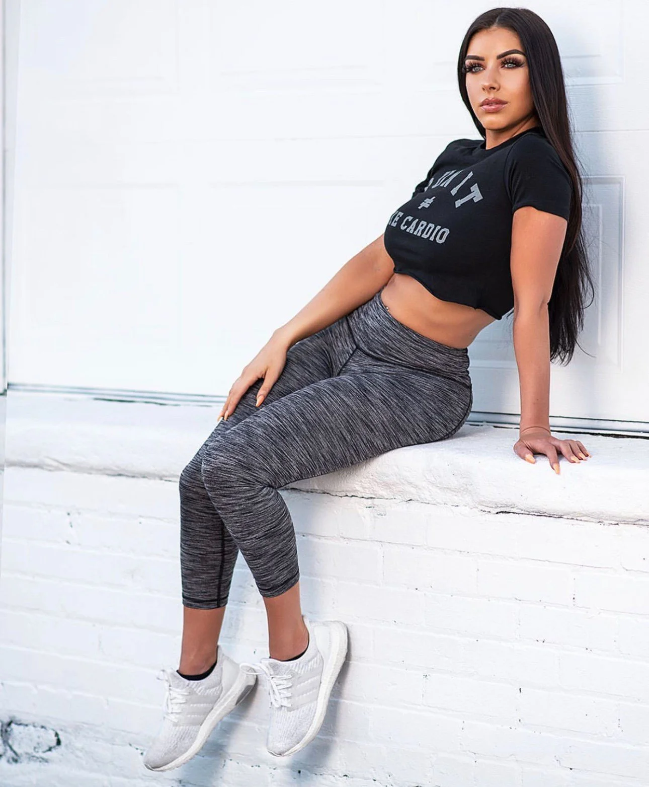 A woman with long dark hair and athletic clothing sitting on a white windowsill against a white brick wall.