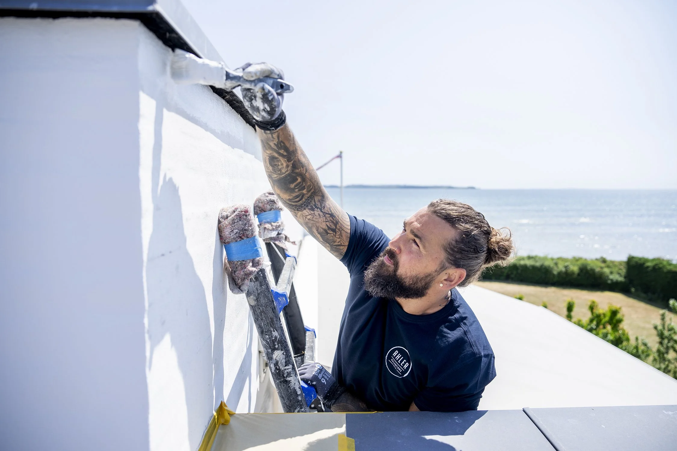 Maler Ahler fra Aarhus arbejder på en udendørs hvid væg, bruger værktøj og beskyttelseshandsker, med baggrund af hav og blå himmel.