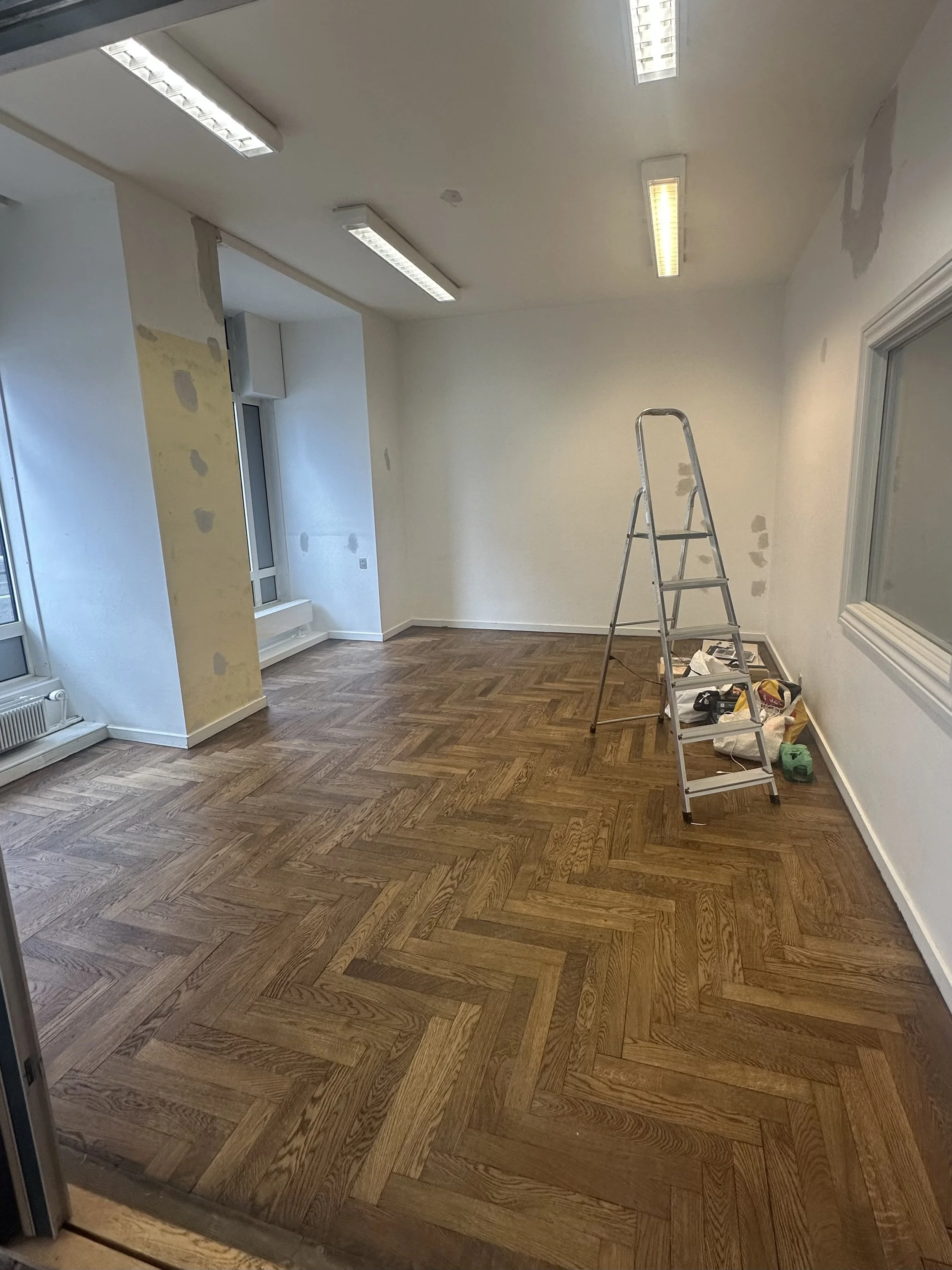 Et rum under renovering med nyt parketgulv, stige, udstyr, og vægge med ufuldstændig opsætning eller maling, udsigten indendørs med vindue og loftlys.