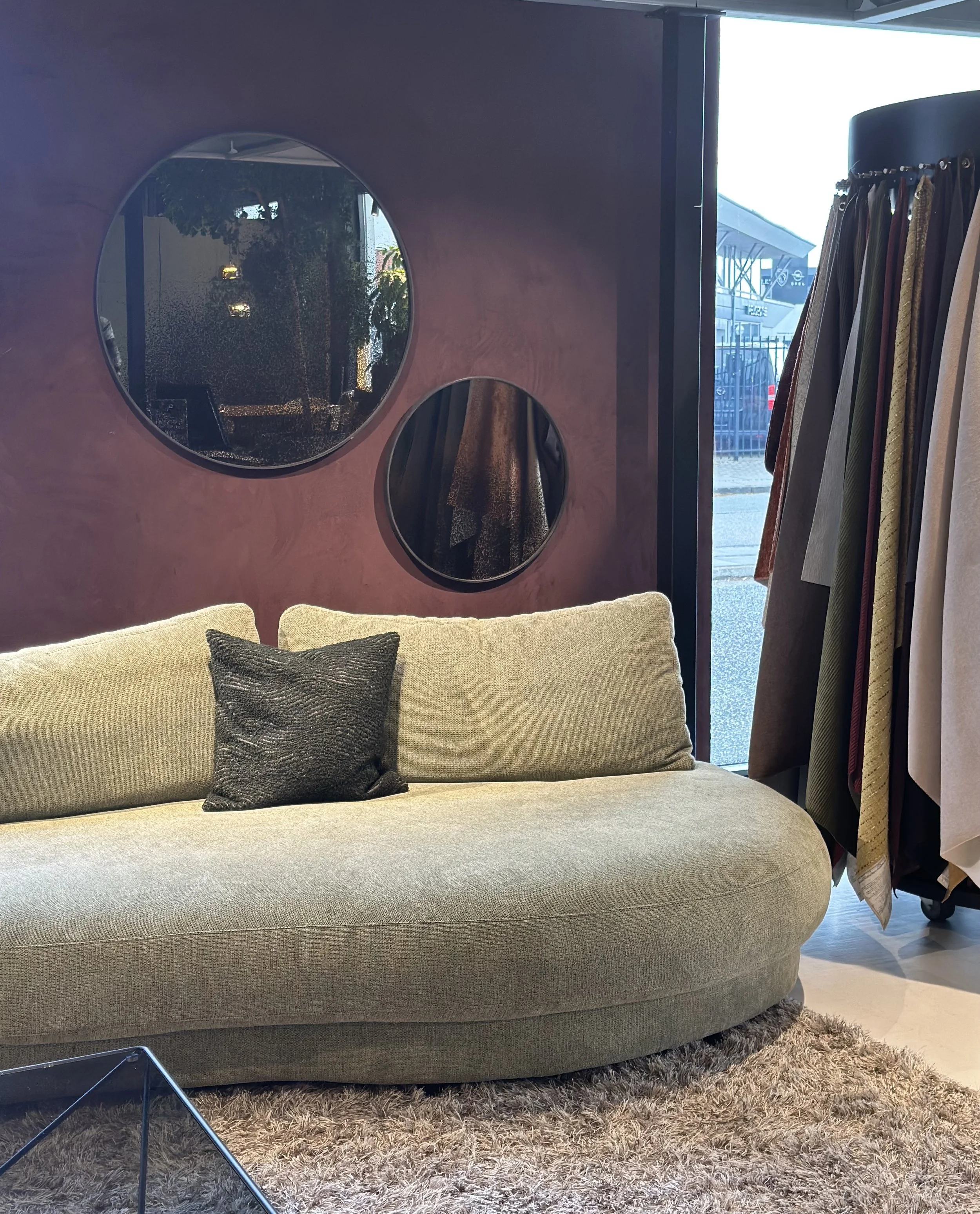 En beige sofa med sort pude foran en lyserød væg med to dekorative runde spejle. Der er tøj hængende på en stang til højre, og et vindue med udsigt til en bygning udendørs.