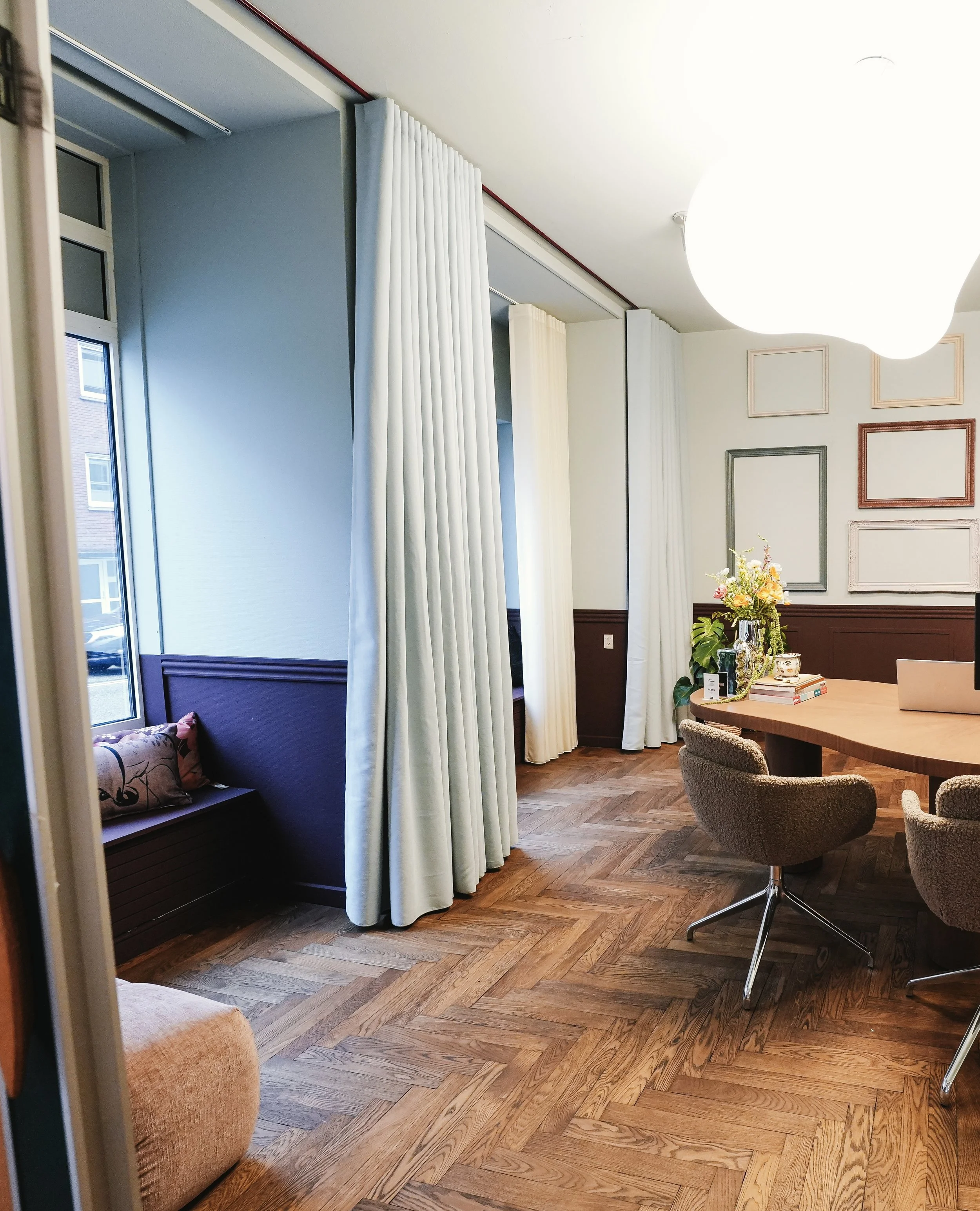 Indendørs billede af et rum med stor hvid lampeskærm, vægdekoration med indrammede fotos, blomsterarrangement på bord, komfortable stole og tæppe foran vindue med hvid gardin.