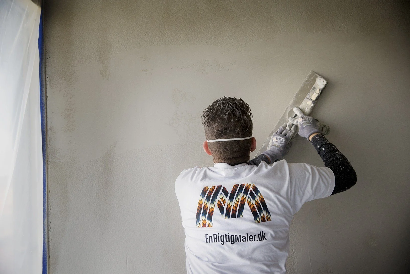 Person retoucher væggen med spartel, iført handsker og en maske, bagpå en hvid t-shirt med EnRigtigMaler.dk logo.