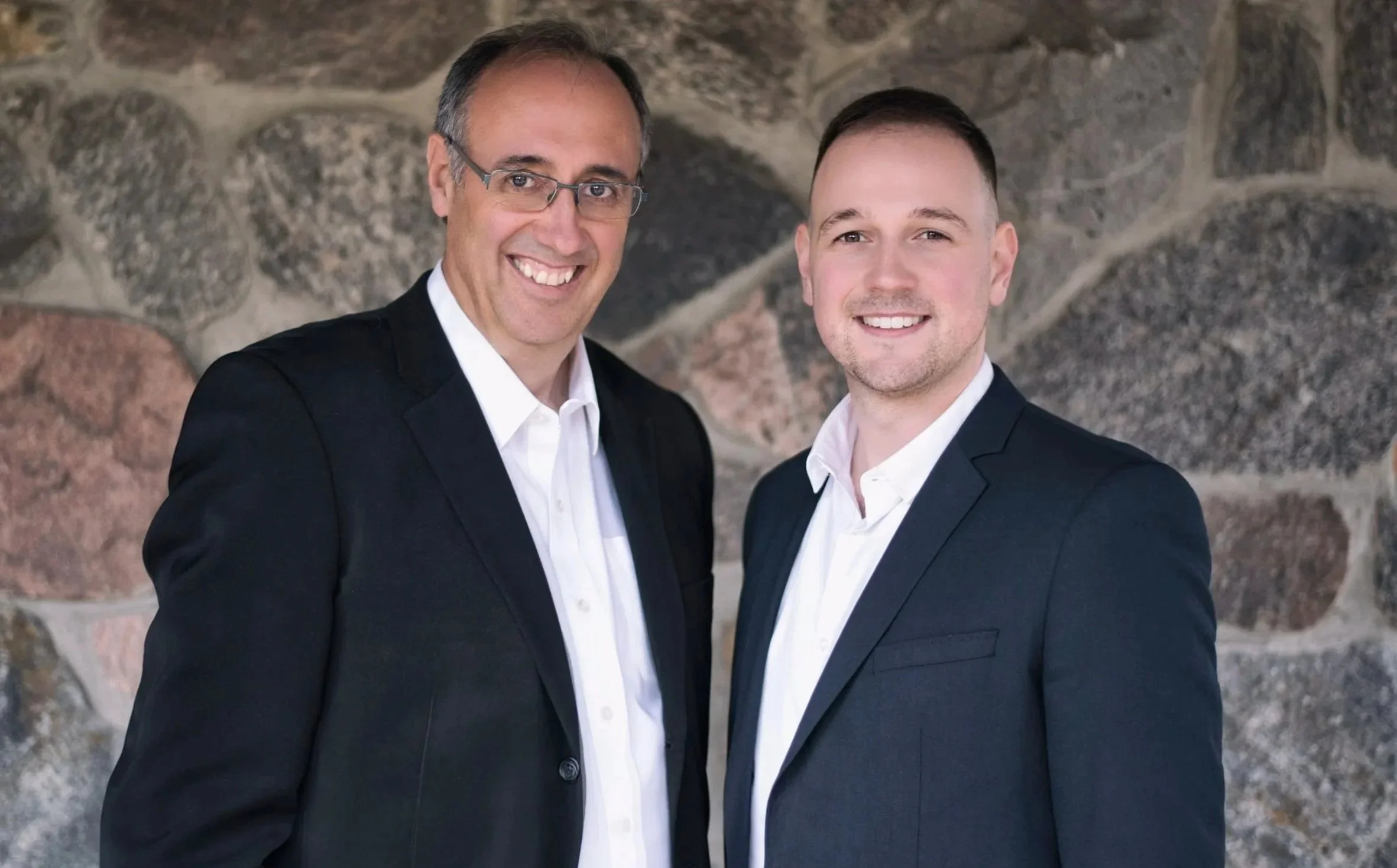 Two men dressed in dark suits and white shirts standing together in front of a stone wall, smiling at the camera.