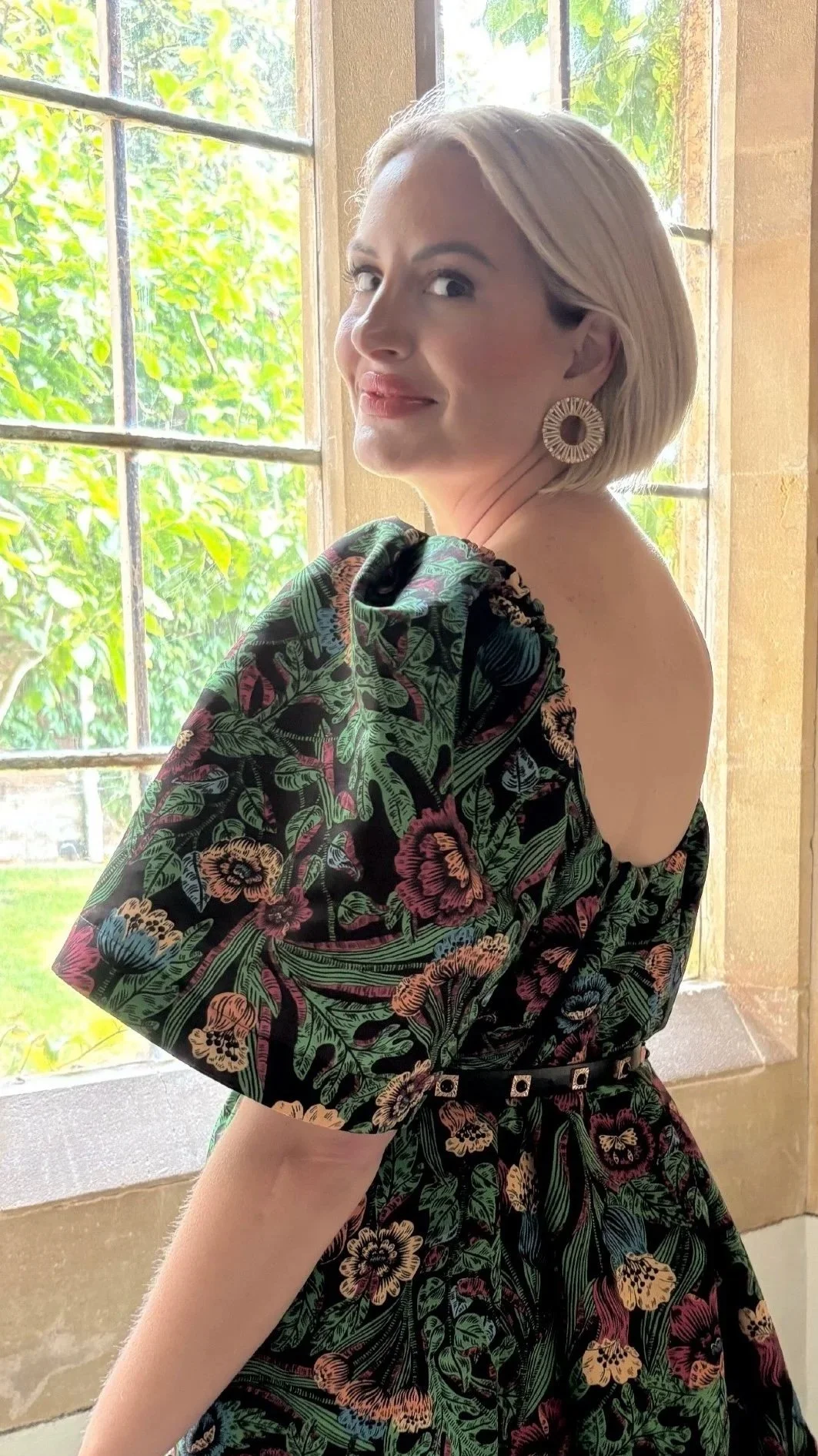 A Bloom Wellness and Aesthetics patient two weeks after her lip flip with Xeomin with short blonde hair and large circular earrings looking over her shoulder inside near a window with green foliage outside.