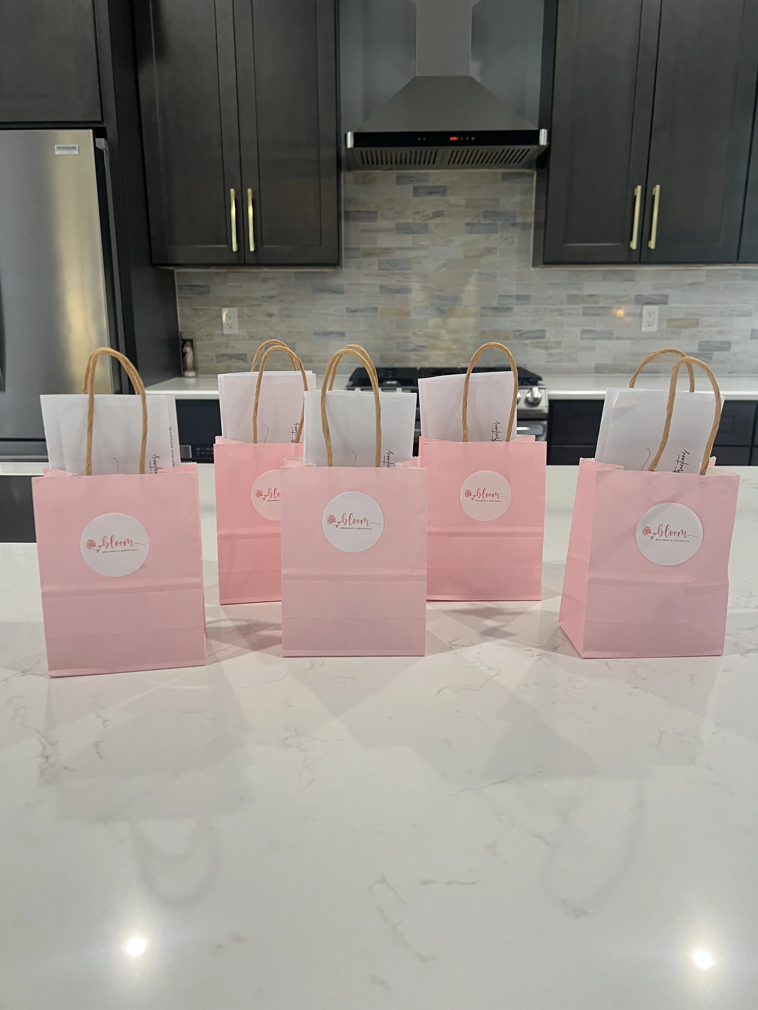 Five pink gift bags with white tissue paper on a white marble kitchen countertop.