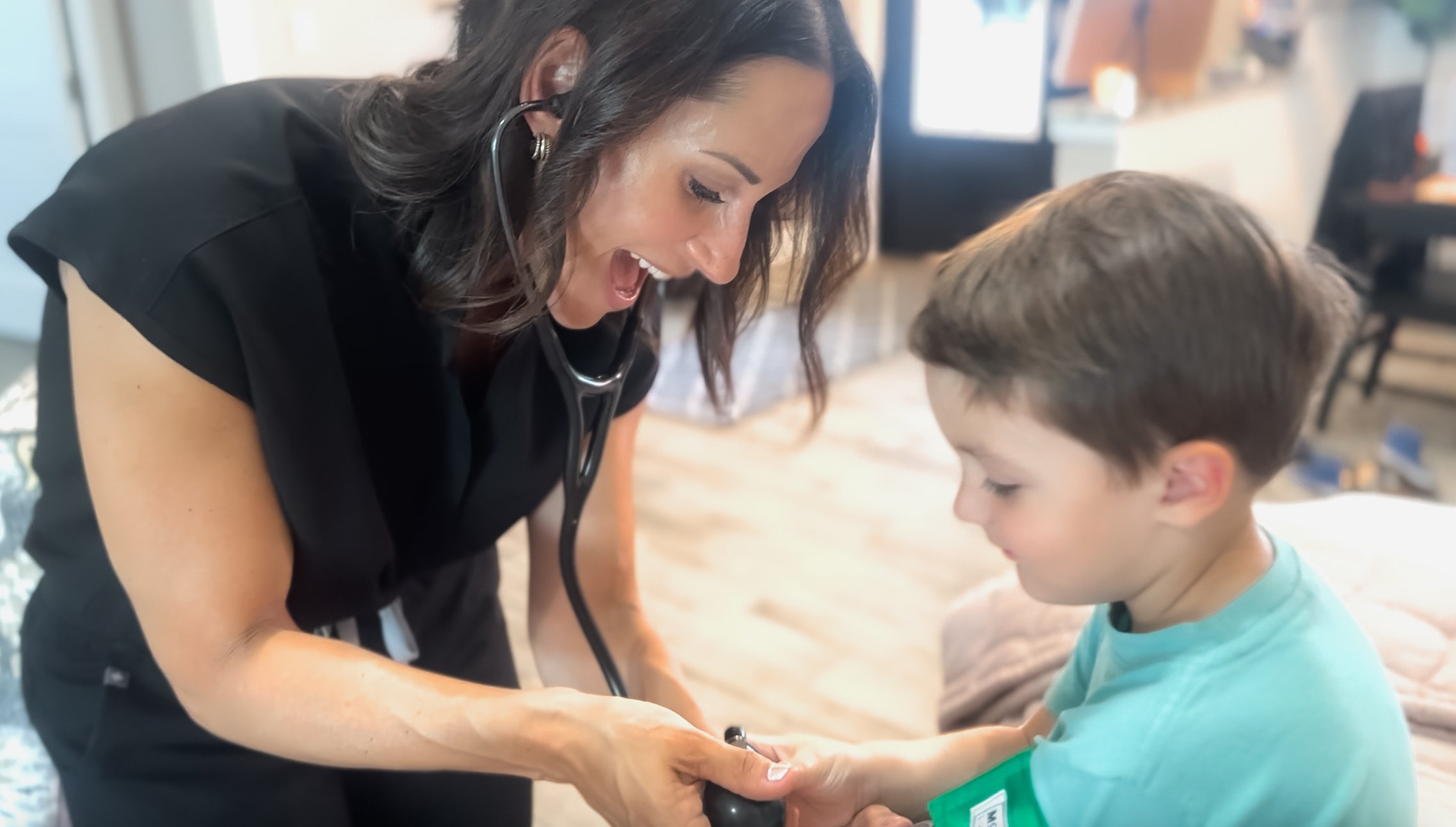 A female healthcare professional, wearing black scrubs and a stethoscope, is smiling and holding a young boy's hand during a check-up or appointment in a medical setting.