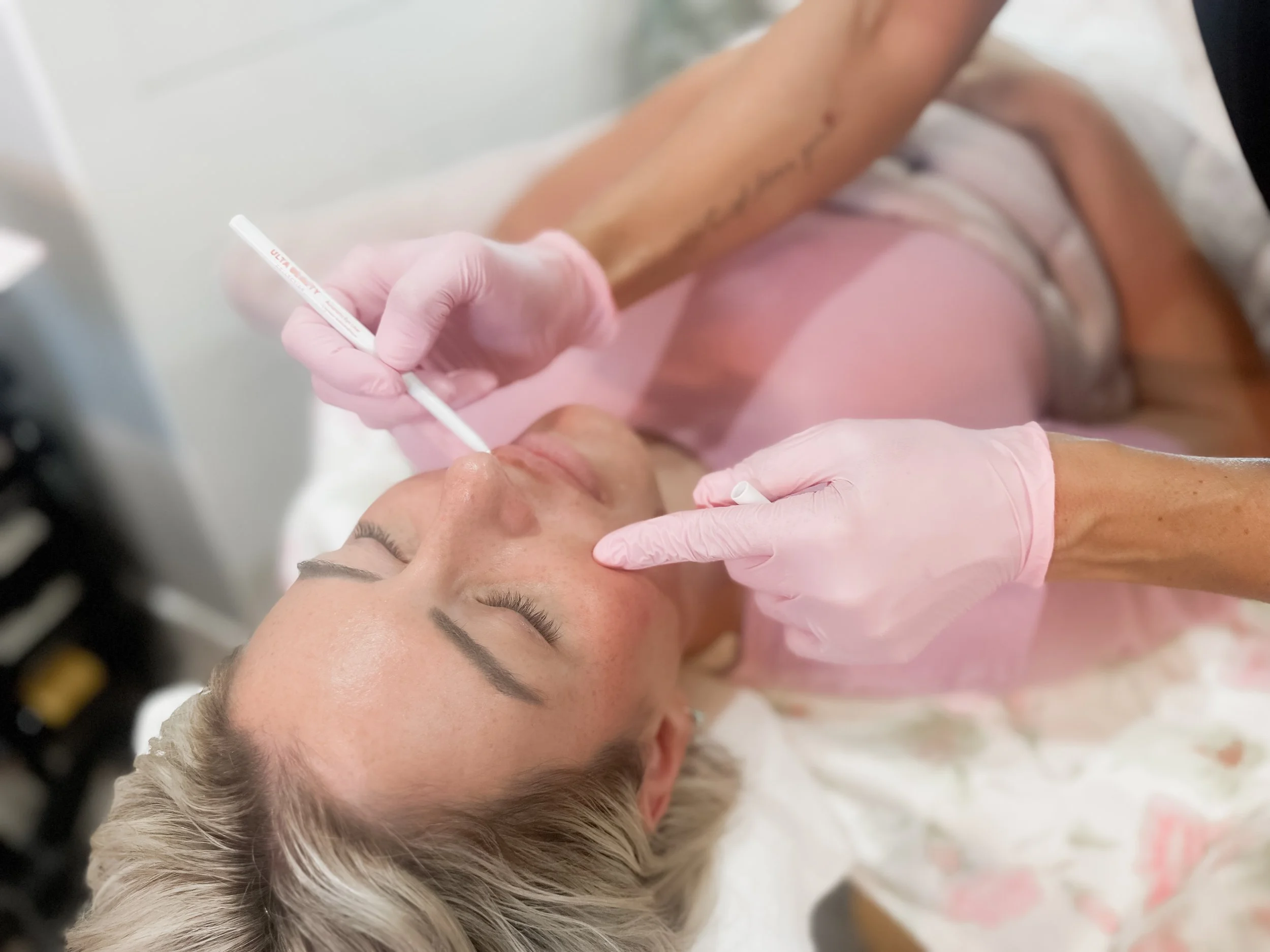 A woman lying on a medical bed receives a cosmetic injection in her forehead from a healthcare professional wearing pink gloves.