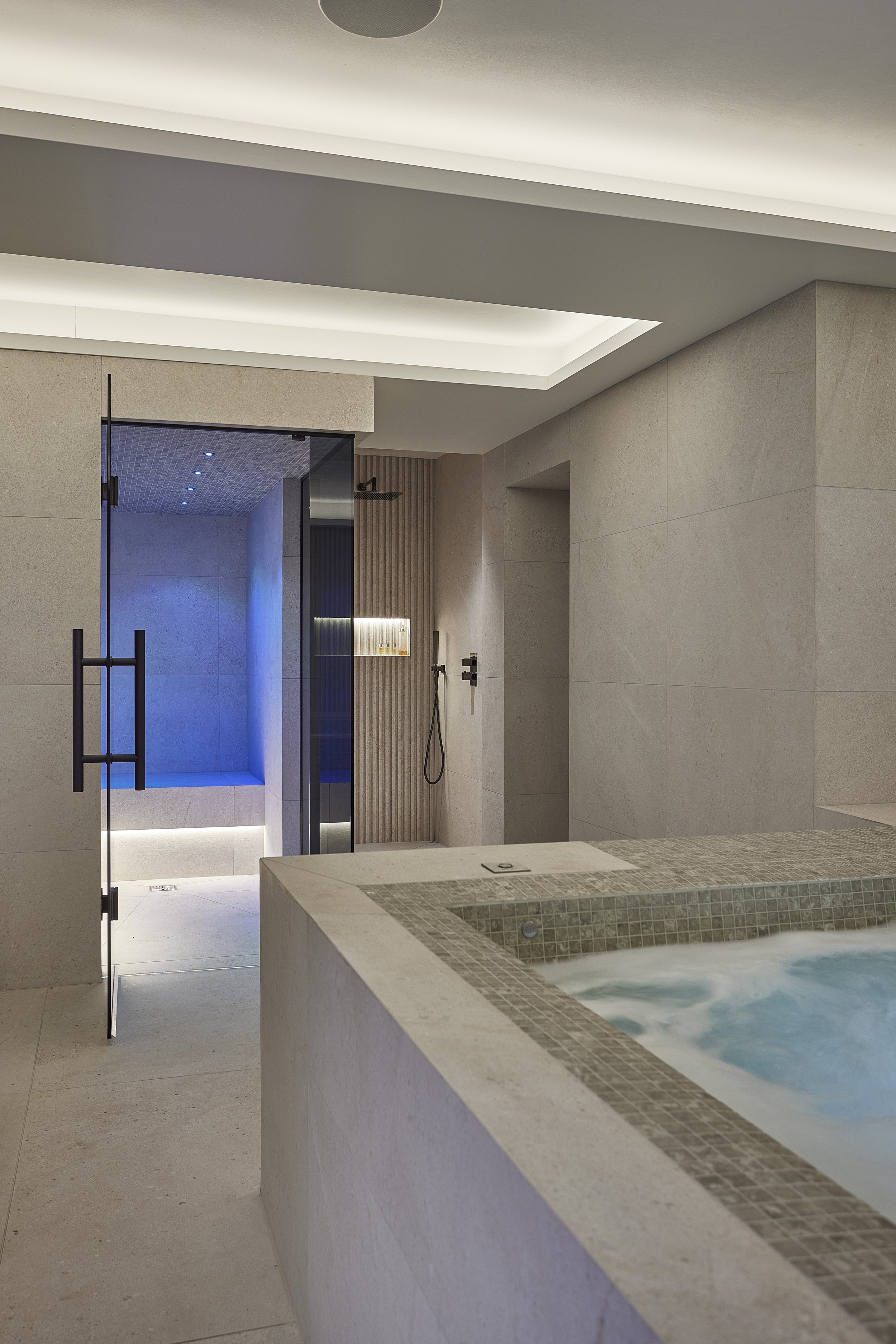 Modern indoor spa with a hot tub in the foreground, and a walk-in shower with a glass door in the background, featuring beige tiles, a ceiling-lit ceiling, and minimalist decor.