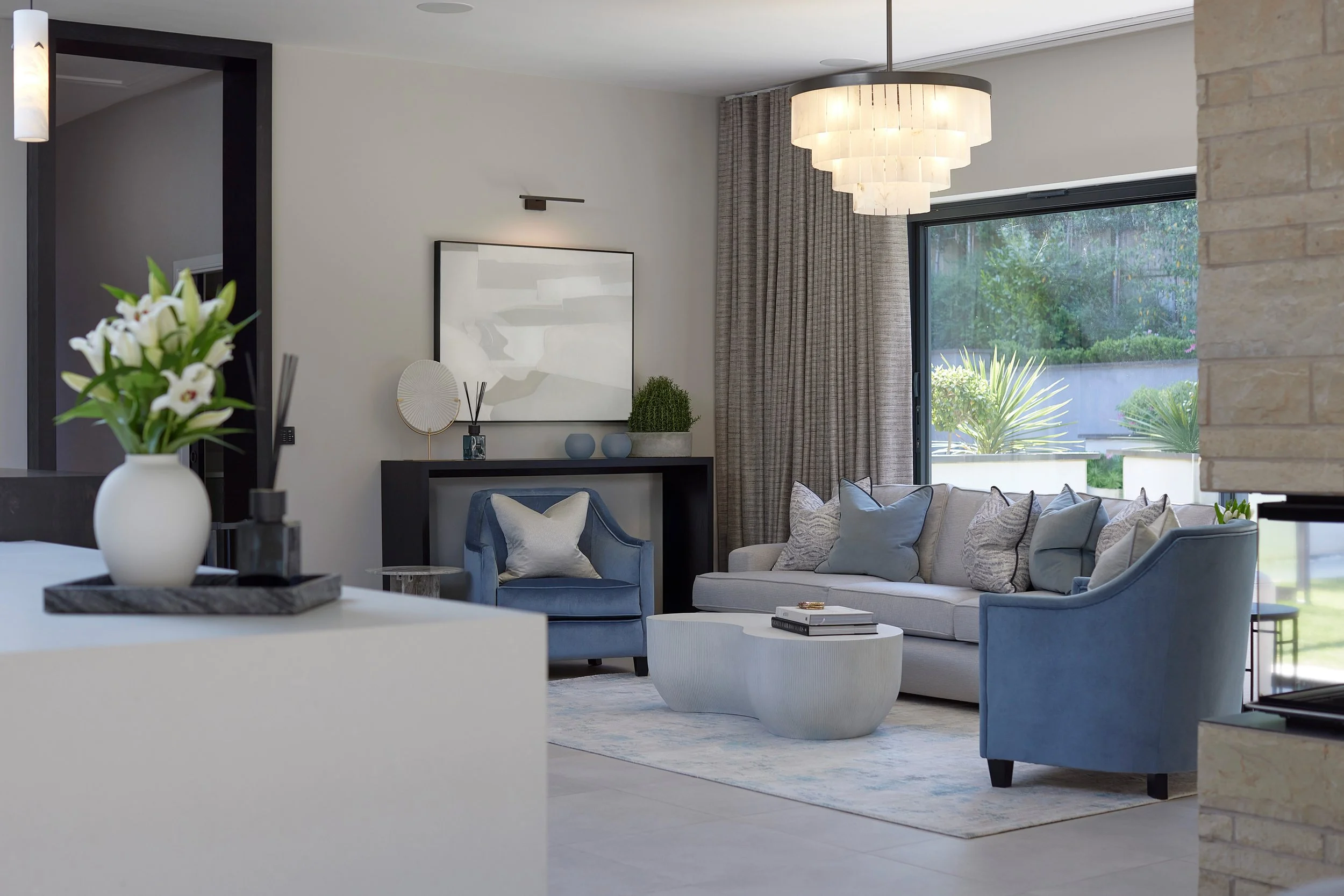 Modern living room with a white sofa, blue armchair, marble coffee table, artwork on the wall, large window with curtains, and a chandelier.