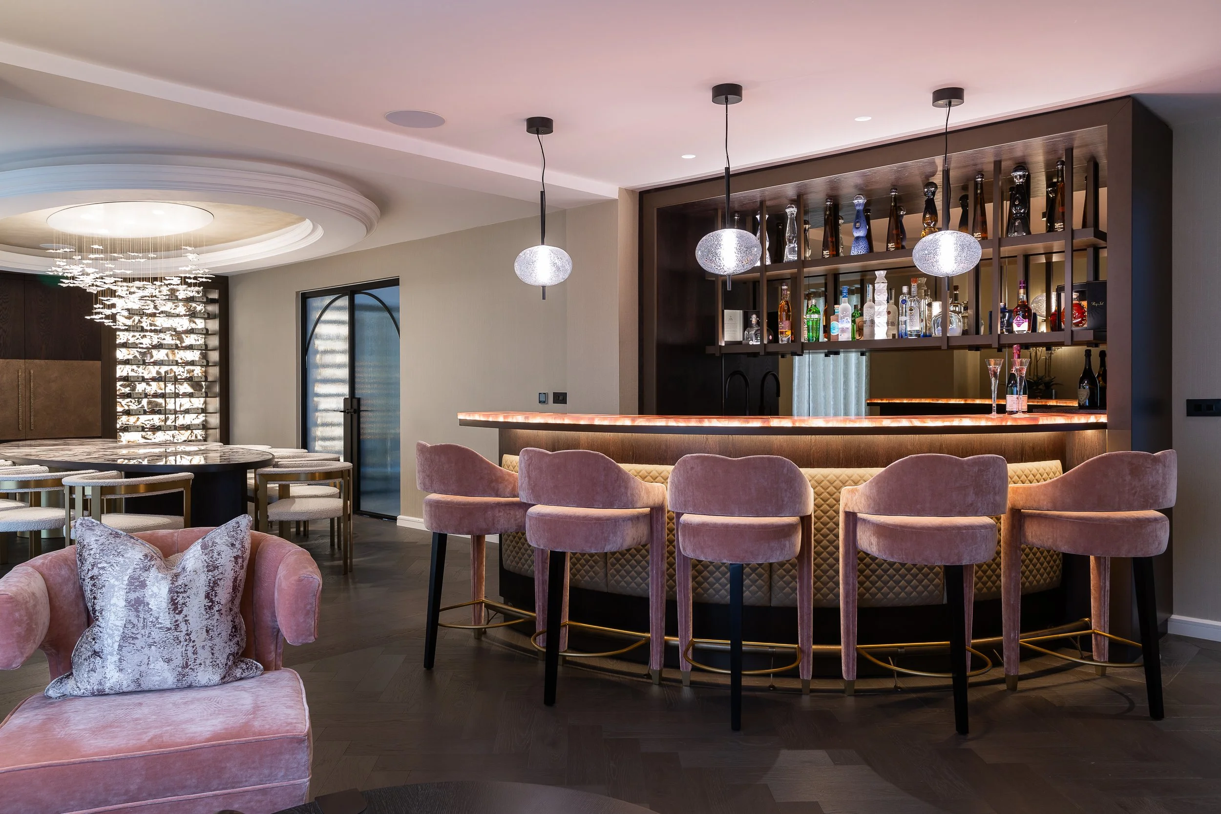 Modern home bar with pink velvet barstools, illuminated counter, and a variety of liquor bottles on dark wooden shelves, with overhead pendant lights.
