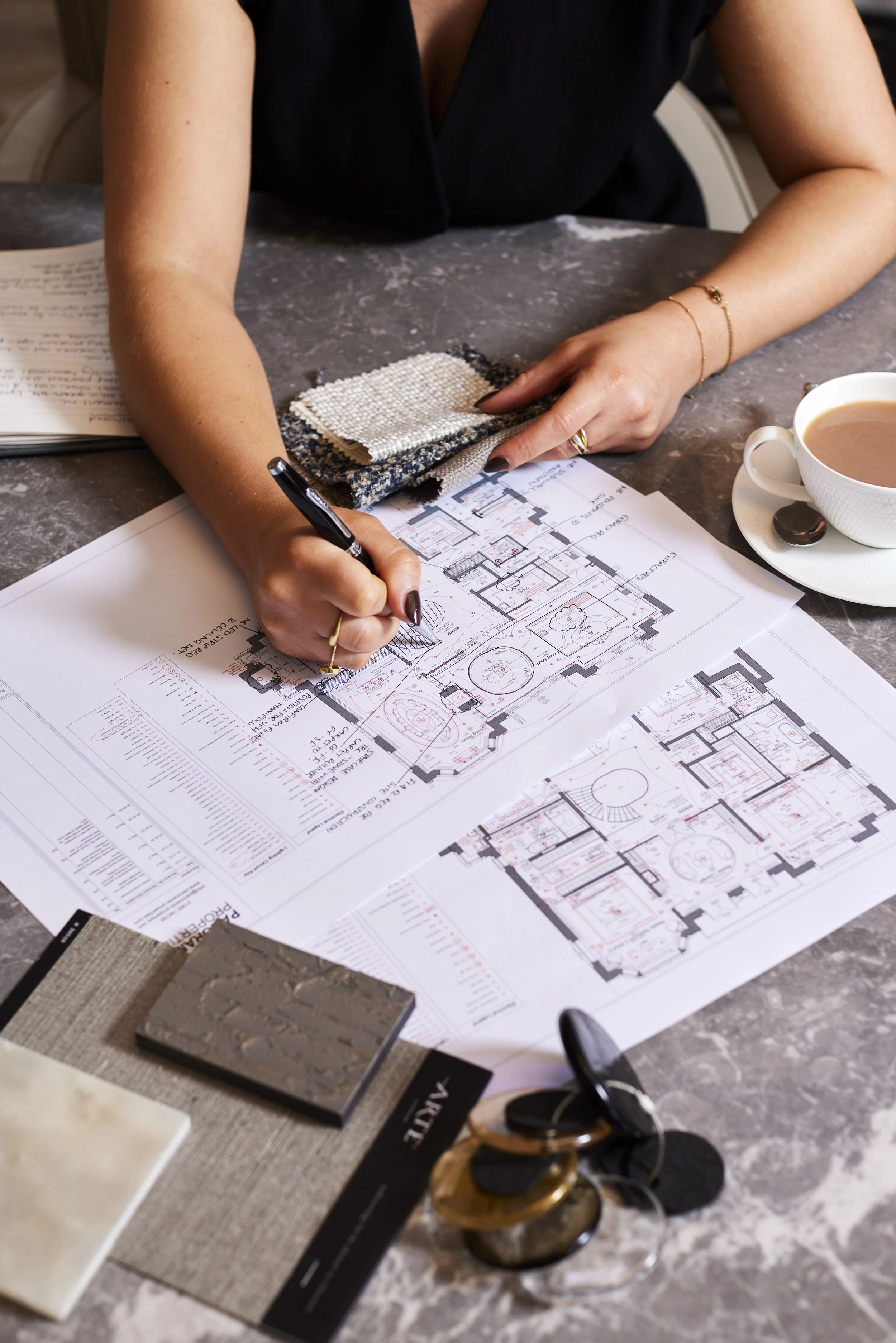 Person working on an architectural floor plan with fabric and material samples, cup of coffee, and design tools on a gray marble table.