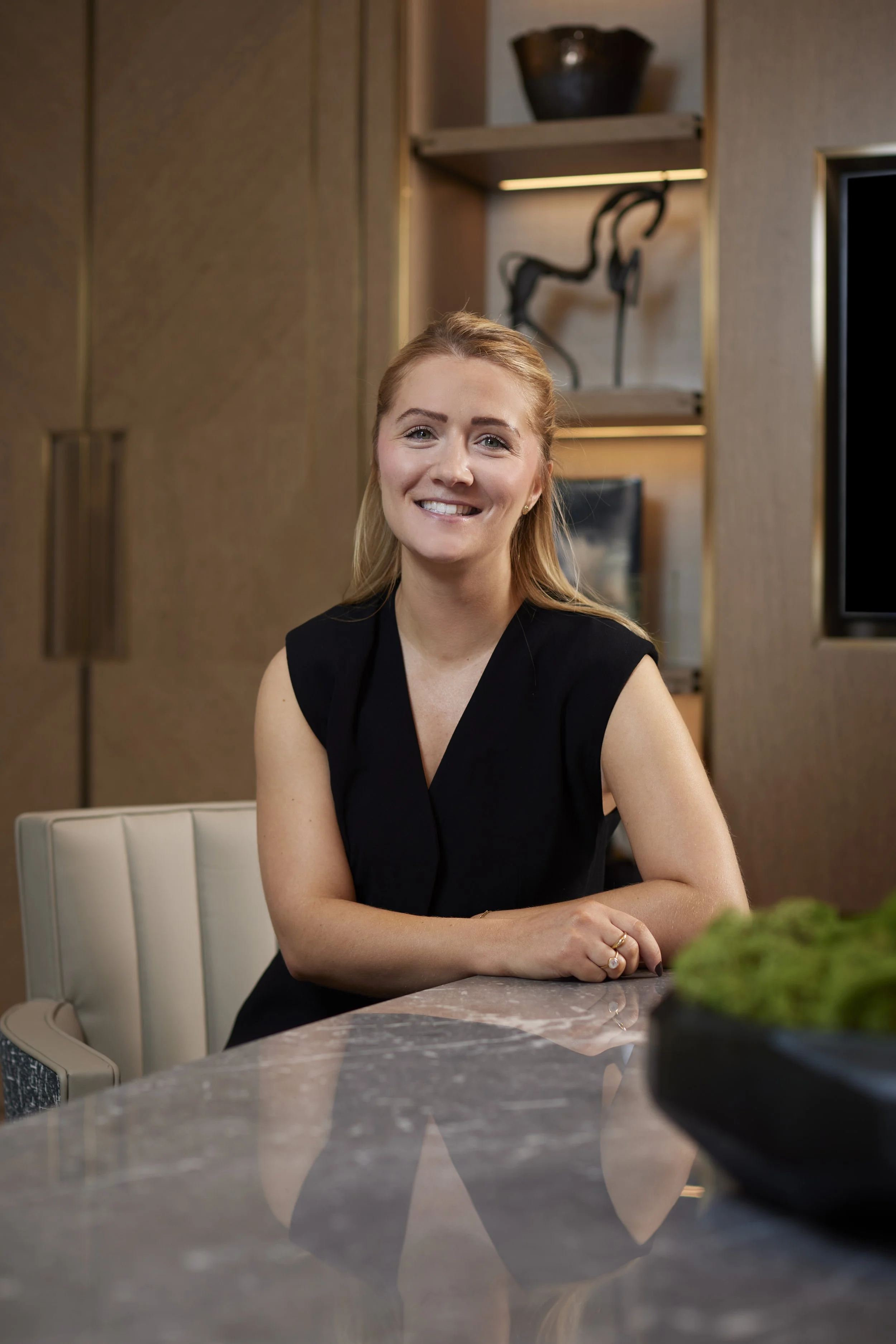 A smiling woman with long blonde hair wearing a black sleeveless top, sitting at a marble table in a modern, well-lit room.