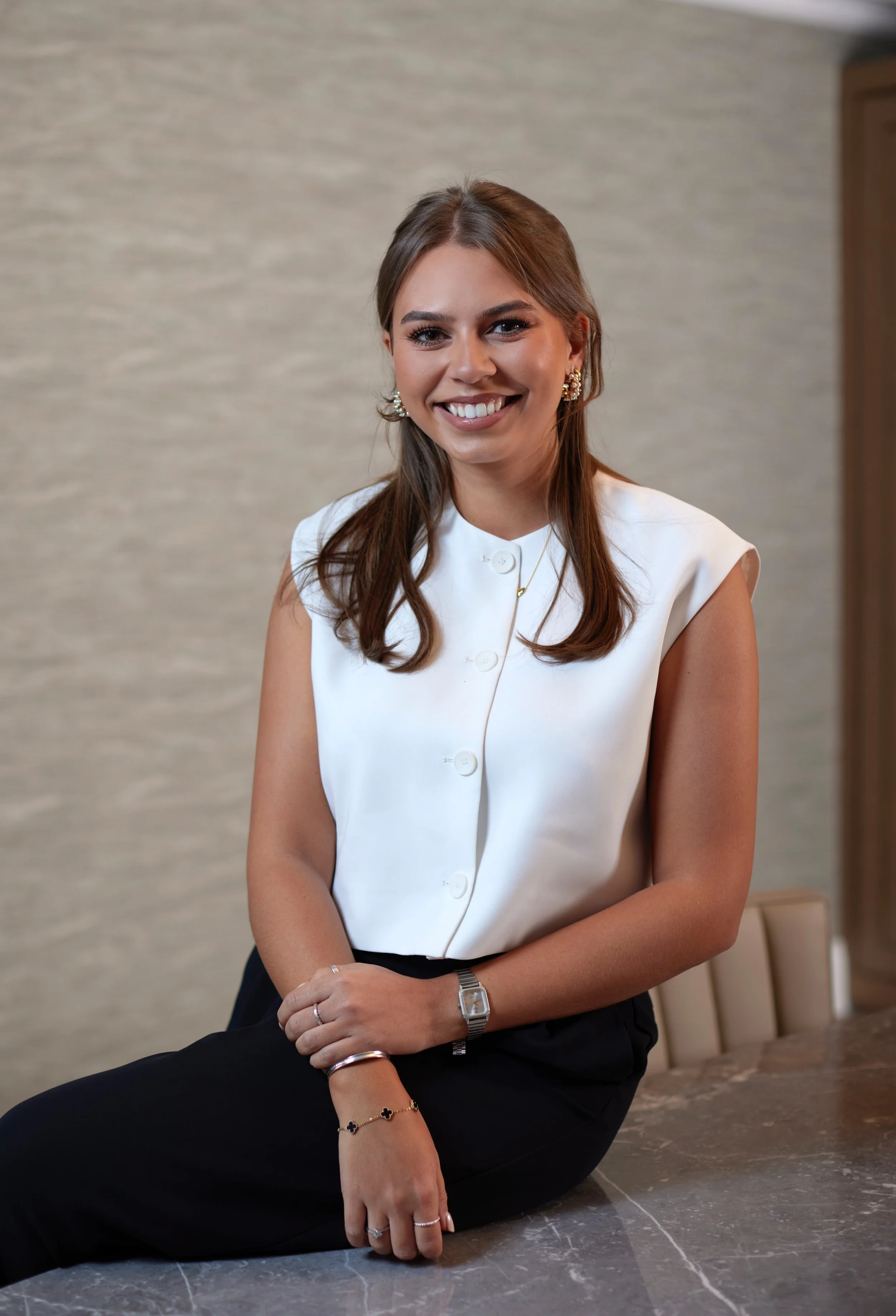 A young woman with shoulder-length brown hair, smiling, wearing a white sleeveless button-up top, black pants, and jewelry, sitting on a marble table in an indoor setting.