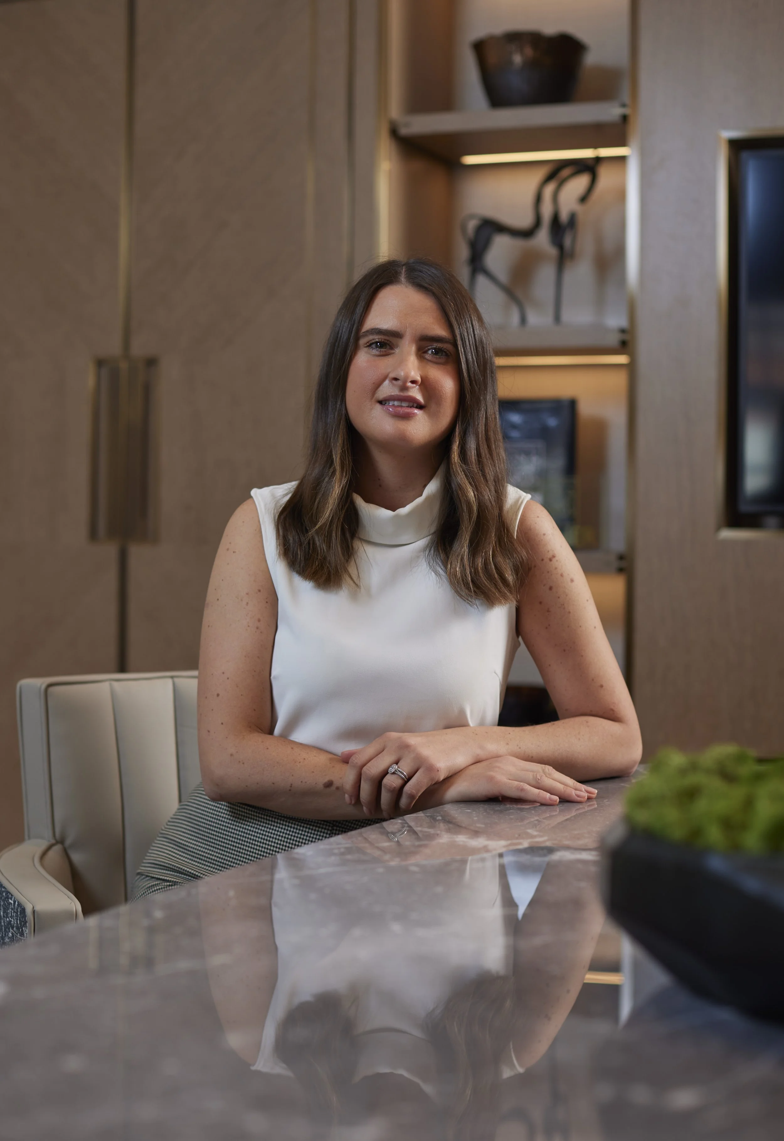 A woman with brown hair wearing a white sleeveless top sitting at a marble table in a modern room with wooden accents and decorative sculptures in the background.