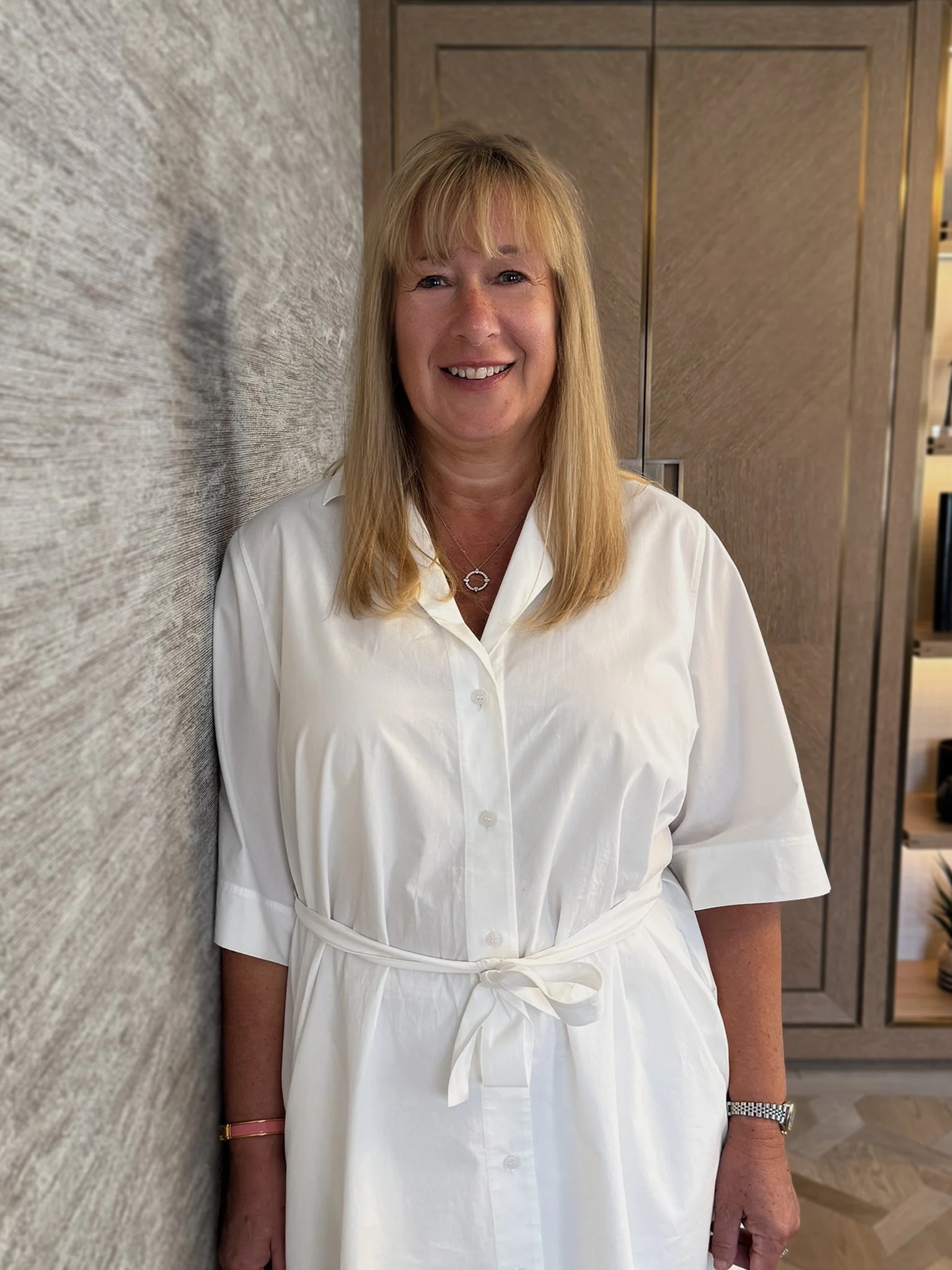 A smiling woman with blonde hair, wearing a white dress, standing inside a modern room with a textured gray wall and wooden panels behind her.
