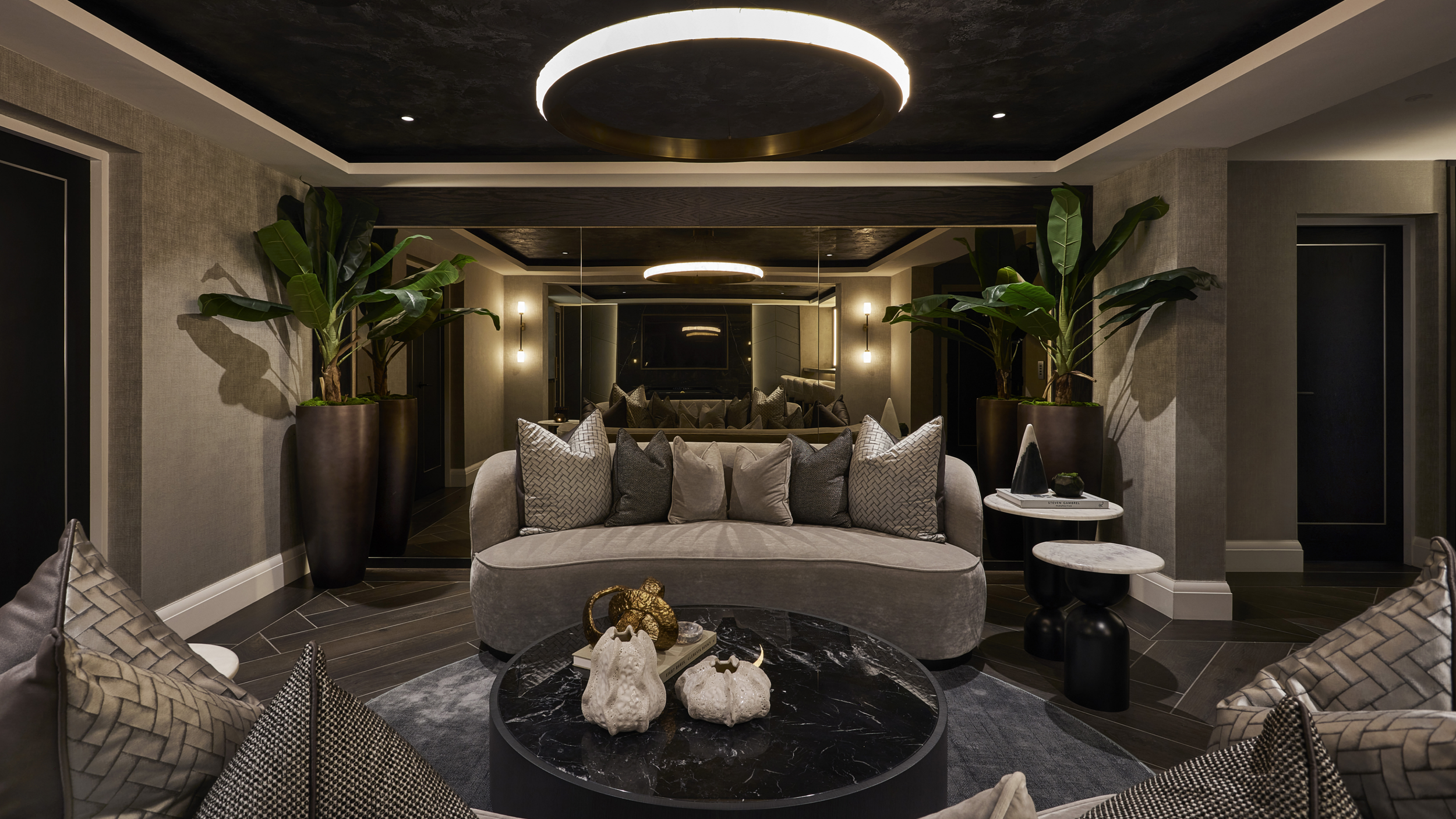 Elegant living room with a beige sofa, multiple throw pillows, and modern decor, including potted plants, a black marble coffee table with sculptures, and mirrored walls.