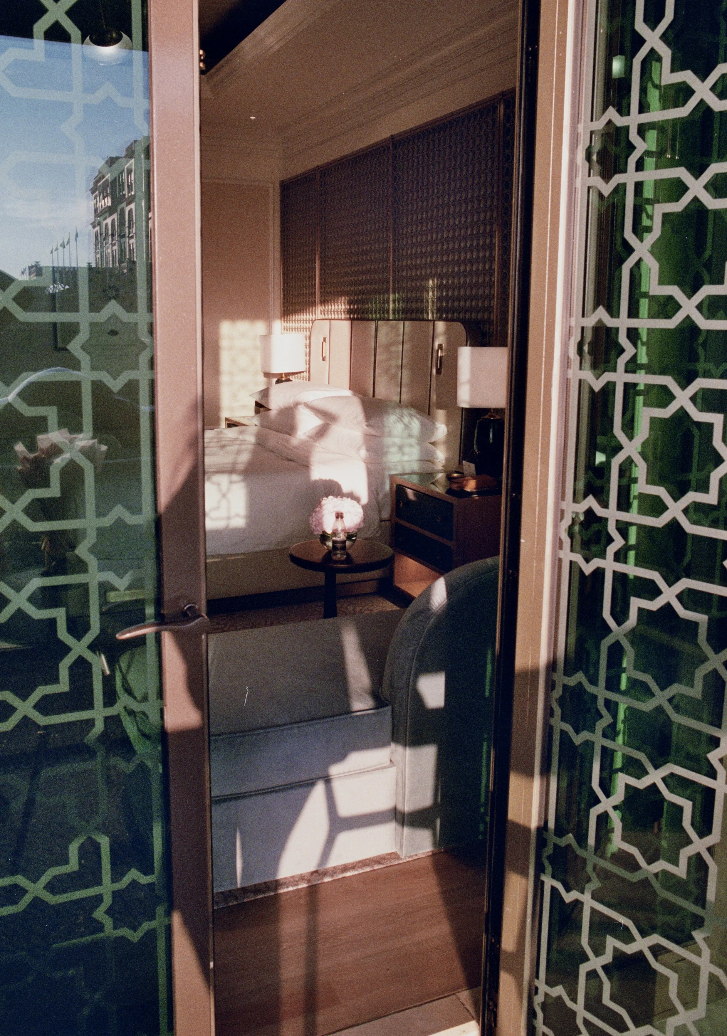 View of a hotel room through a glass door with geometric patterns, showing a bed with pillows, bedside lamps, a small table with a flower arrangement, and a chair. Sunlight casts shadows inside the room.