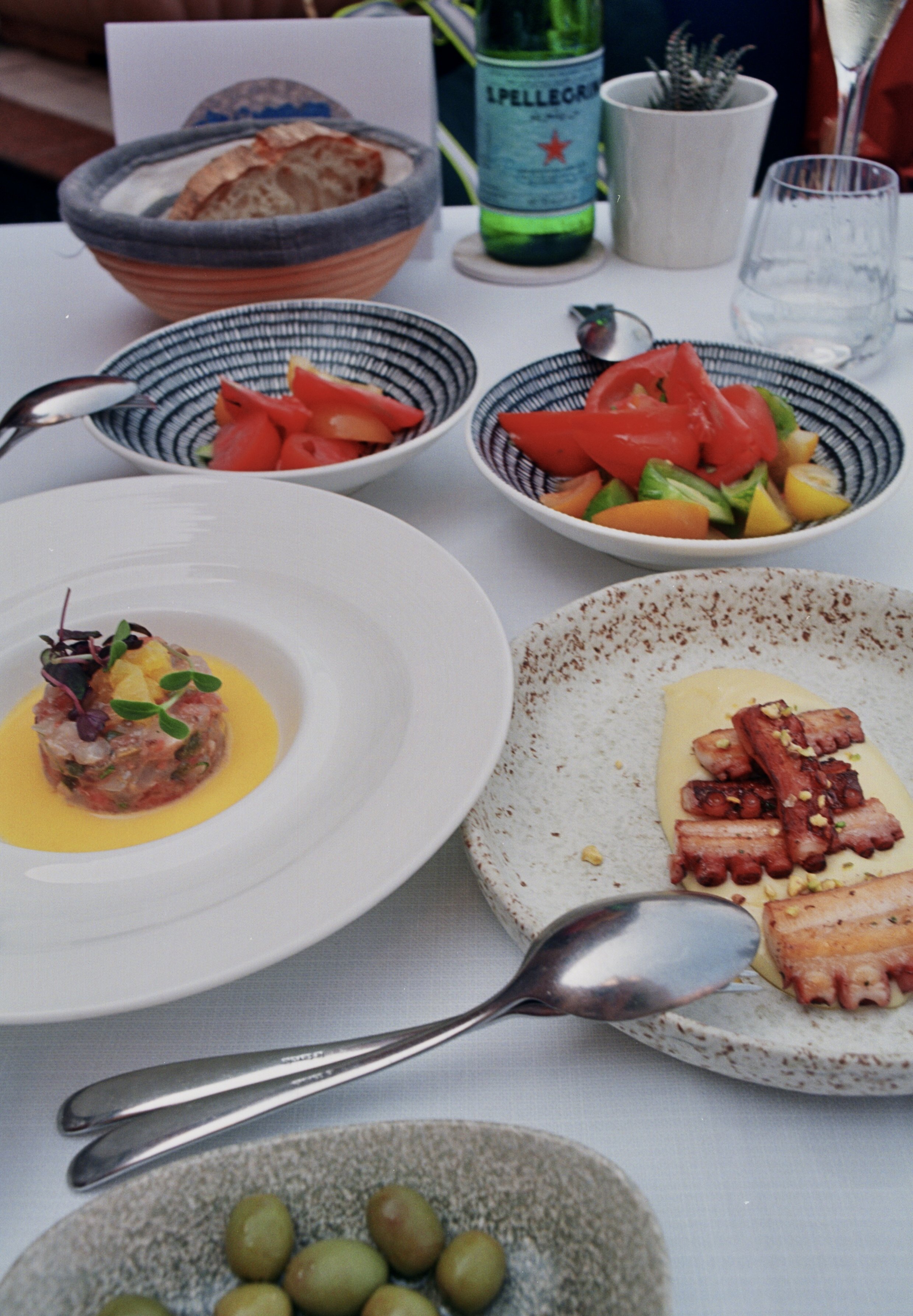 A variety of dishes on a white table, including tomato salads, grilled meats, bread, a bowl of green olives, and bottled water, set in a dining setting.