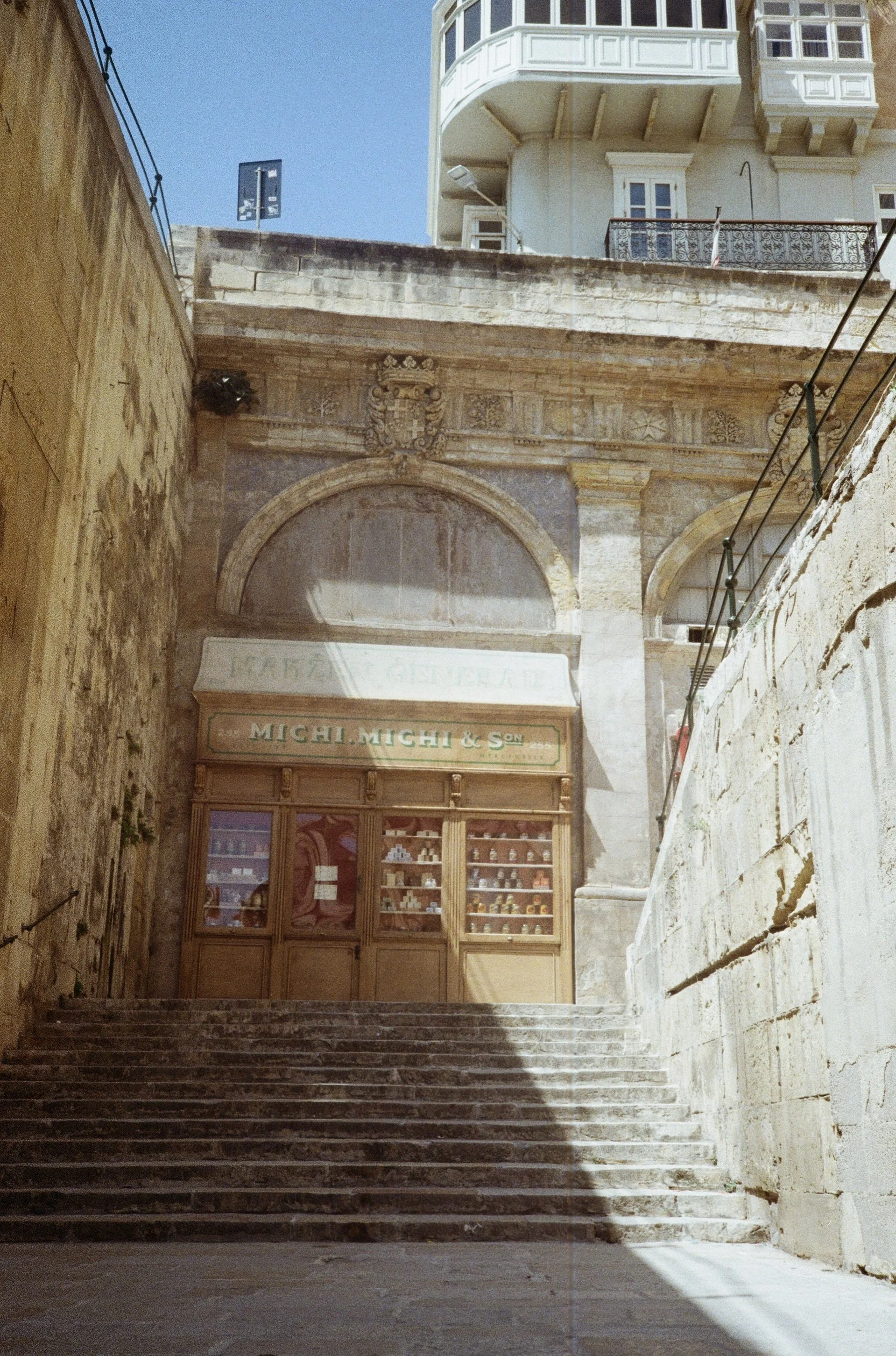Stone staircase leading up to a shop named 'Mich.Michi & S' with display windows, old stone walls on sides, and a historic building with decorative architecture in the background under a clear blue sky.