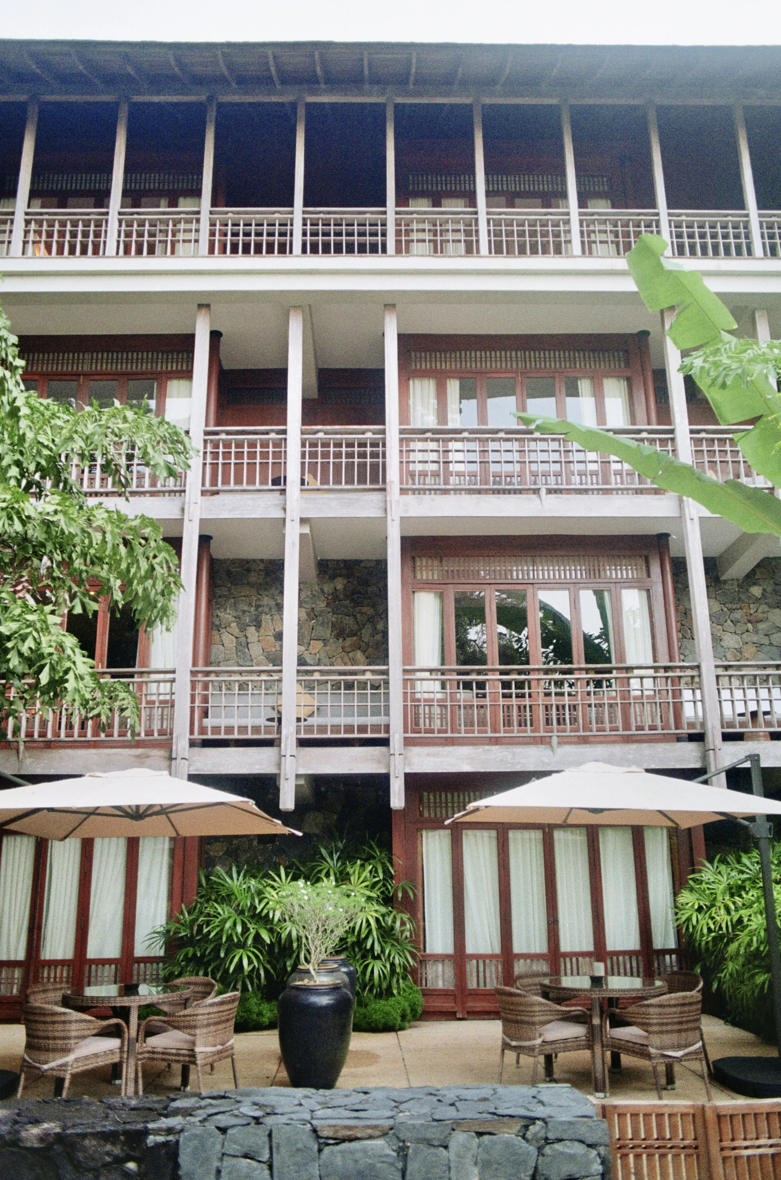 A multi-story building with balconies, surrounded by lush green plants, and outdoor seating with umbrellas in the foreground.