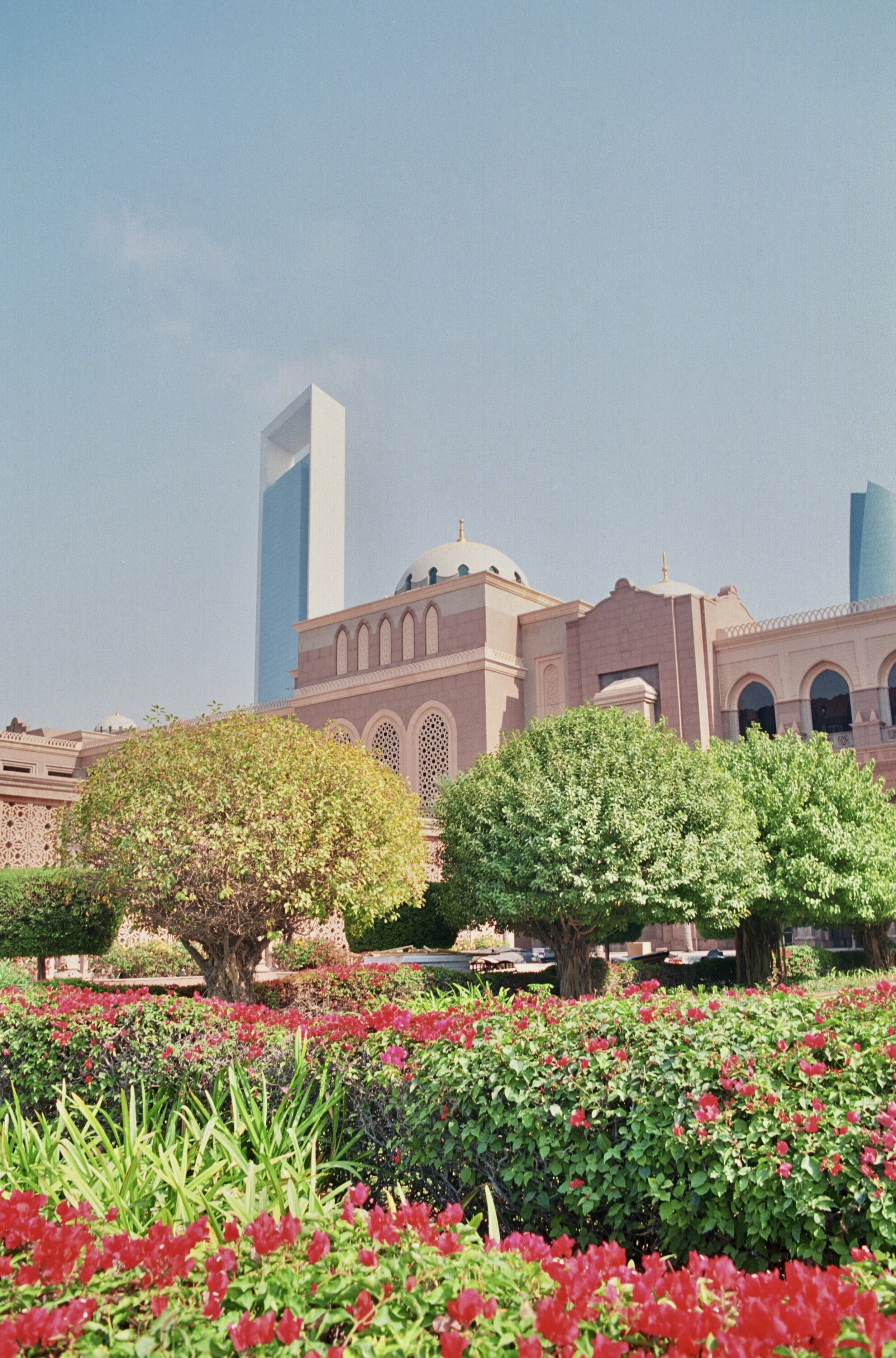 A garden with pink and red flowers, green trees, and a pink Islamic-style building with a dome and arches, against a backdrop of modern skyscrapers and a blue sky.