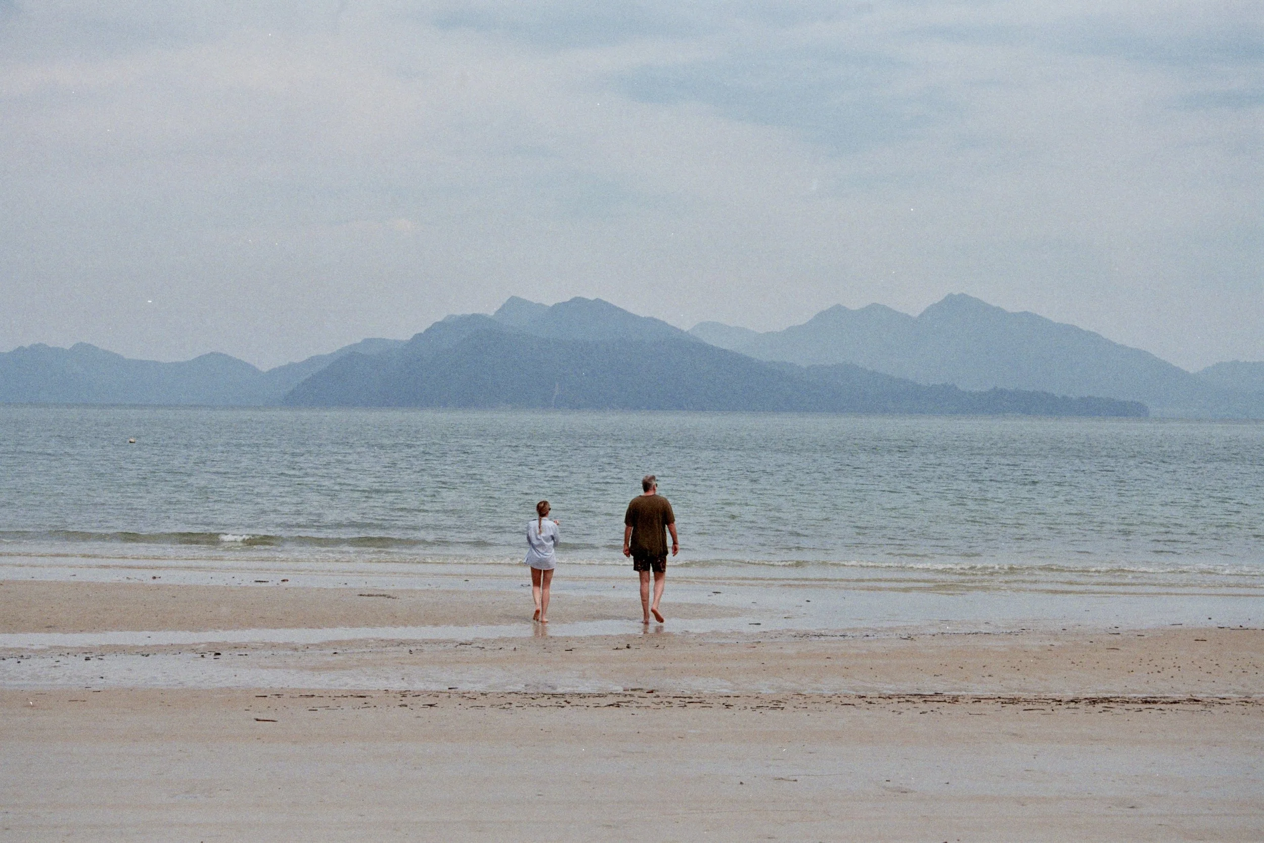 A man and a woman walking along a sandy beach, facing the ocean with mountains in the distance under a cloudy sky.