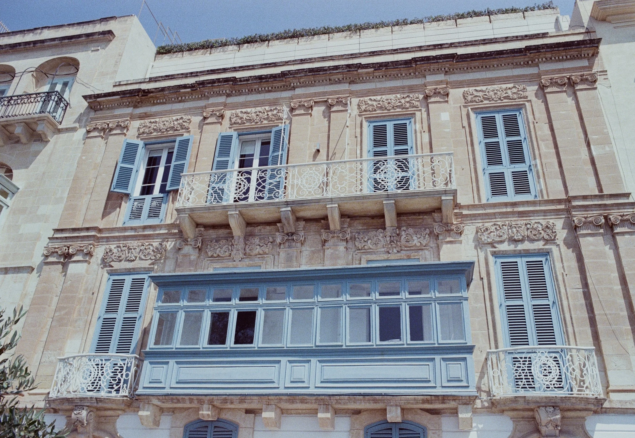 Facade of a historic building with ornate stonework, blue shutters, and balconies with decorative wrought iron railings, under a clear blue sky.