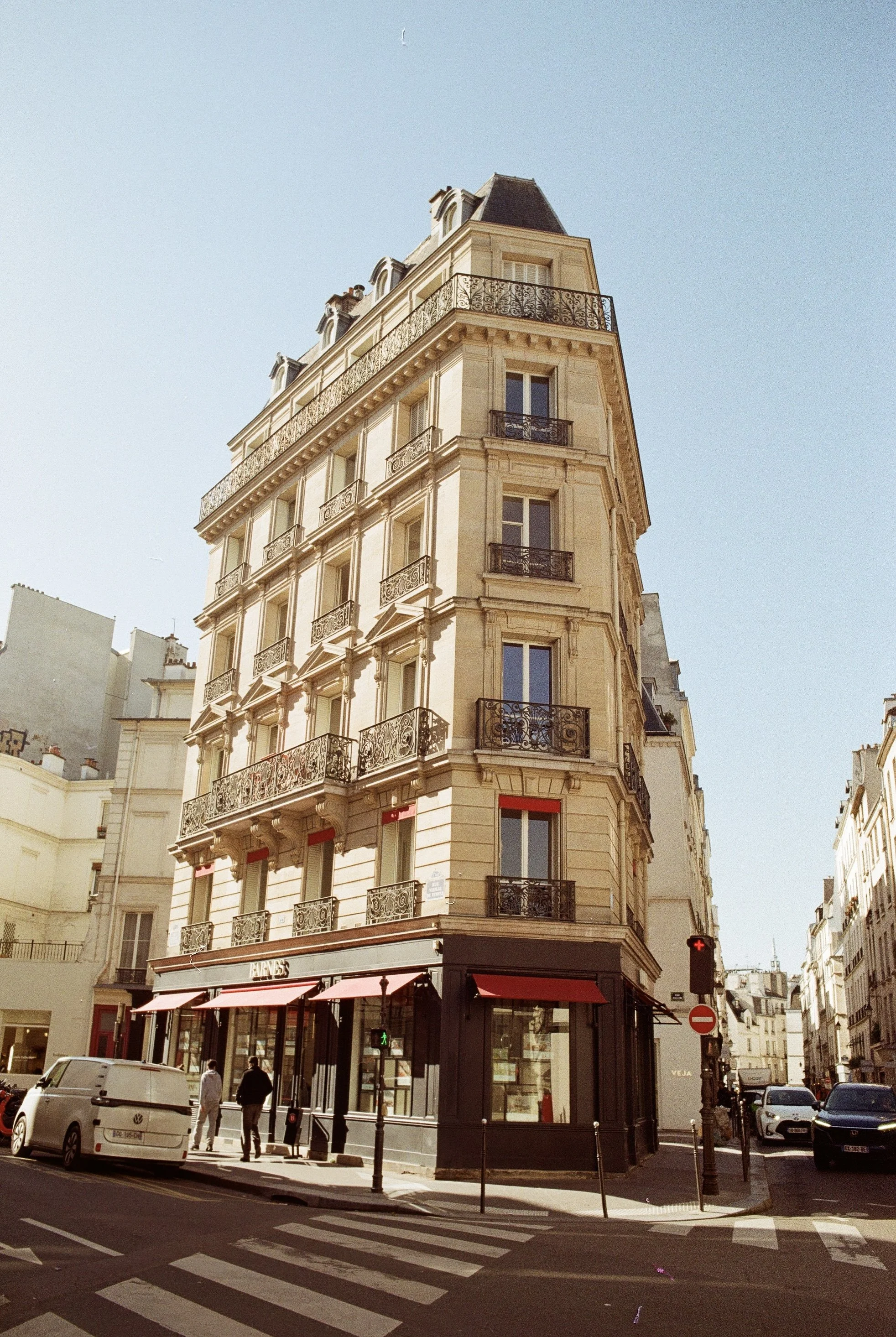 A tall, ornate Parisian building with multiple balconies and black wrought-iron railings, located at an intersection with a few pedestrians and parked cars, under a clear blue sky.