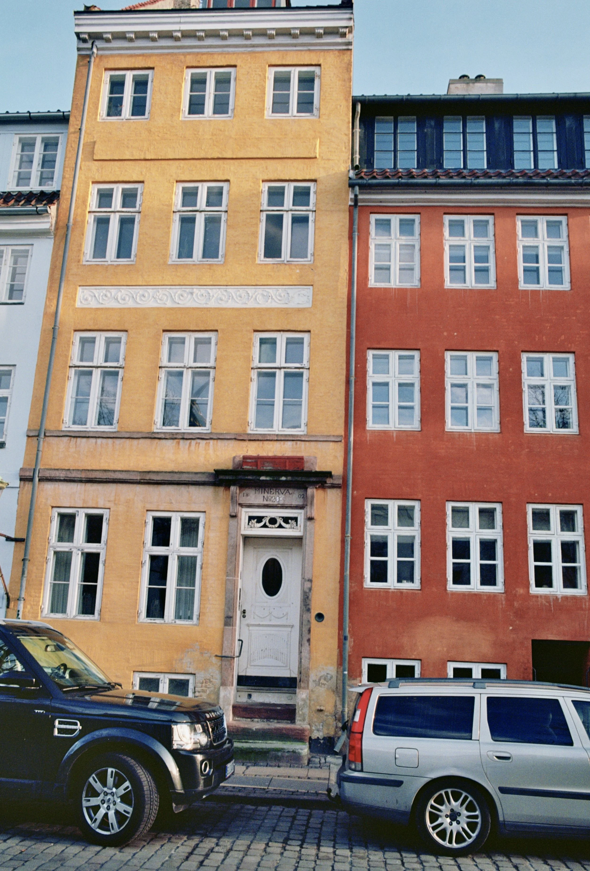 Colorful city buildings with multiple windows, parked cars on cobblestone street.