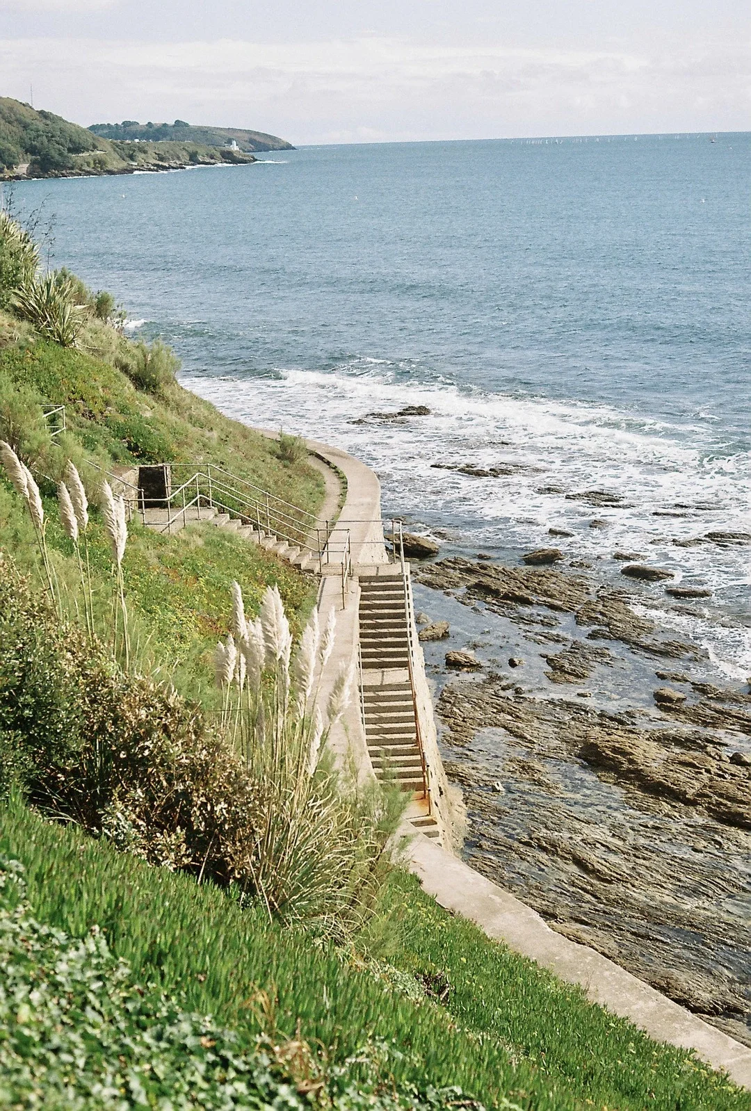 Coastal scene with green plants, stairs leading down to the rocks and ocean, and a distant headland.