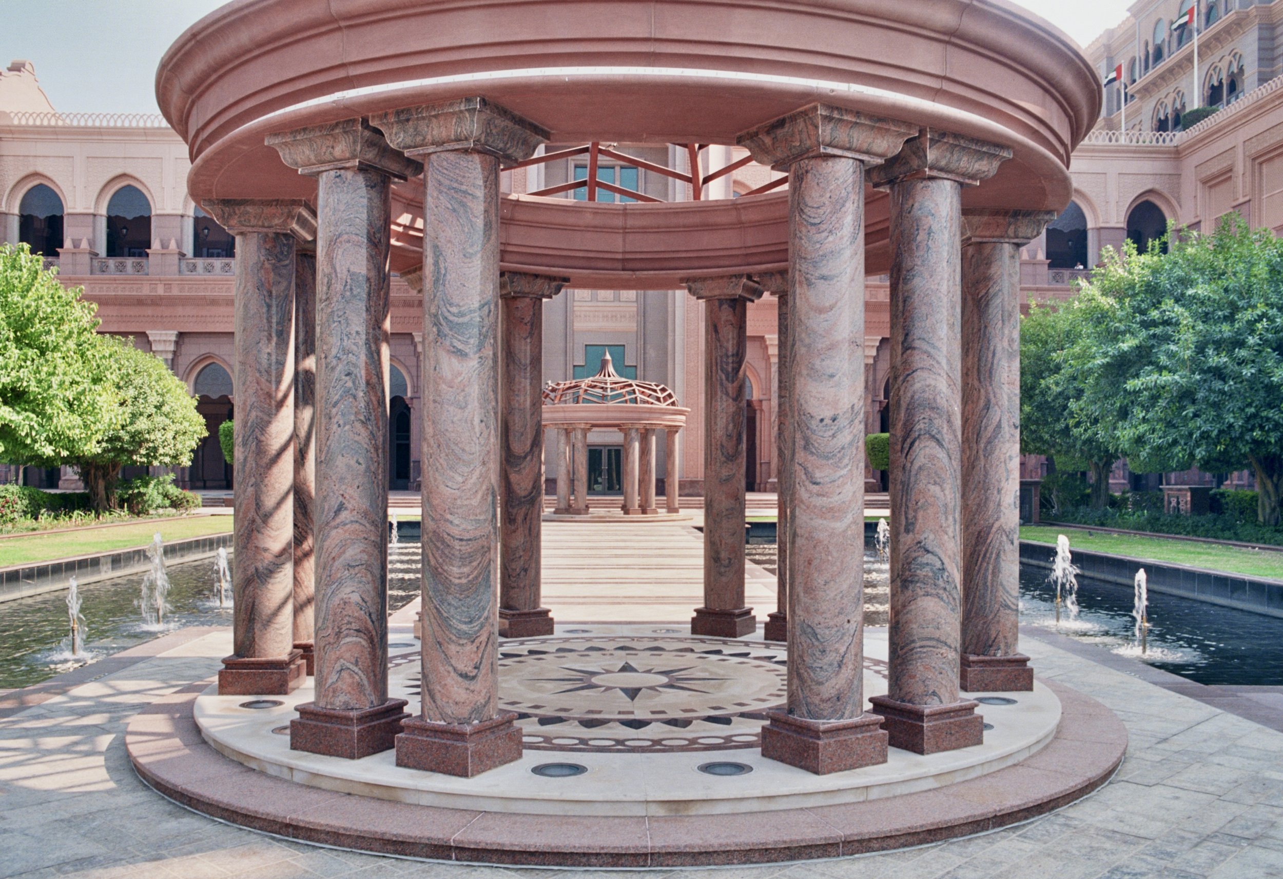 A courtyard with a central marble fountain, surrounded by pink marble columns, reflecting Islamic architectural style with lush green trees and a pink building in the background.